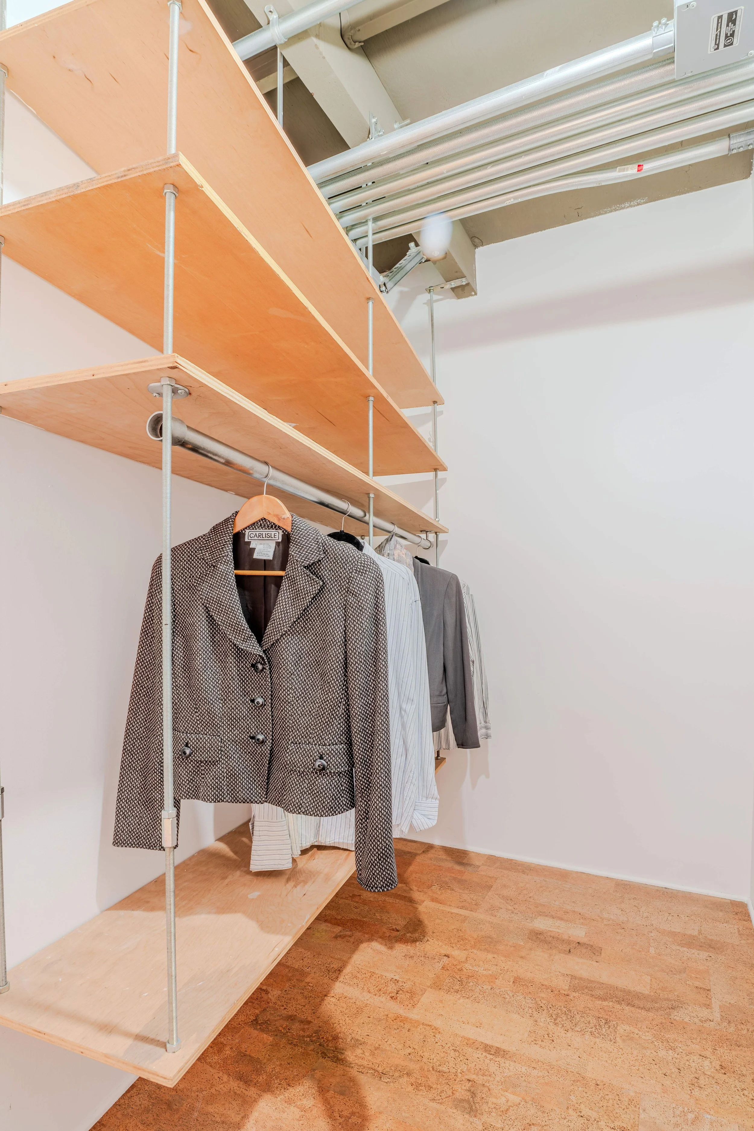 Clothes hanging on a wooden and metal clothing rack in a store or showroom with wooden flooring and a plain white wall.