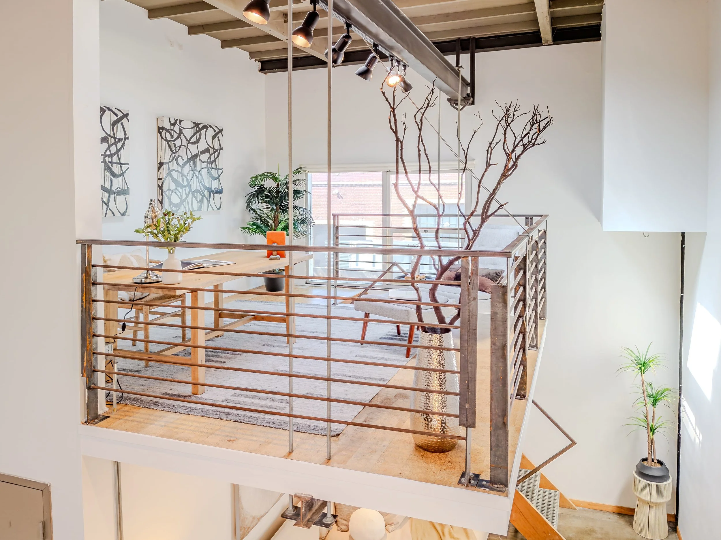 Interior view of a modern loft with a staircase featuring metal railings, a wooden table with a vase of flowers, potted plants, and abstract wall art, illuminated by ceiling spotlights.