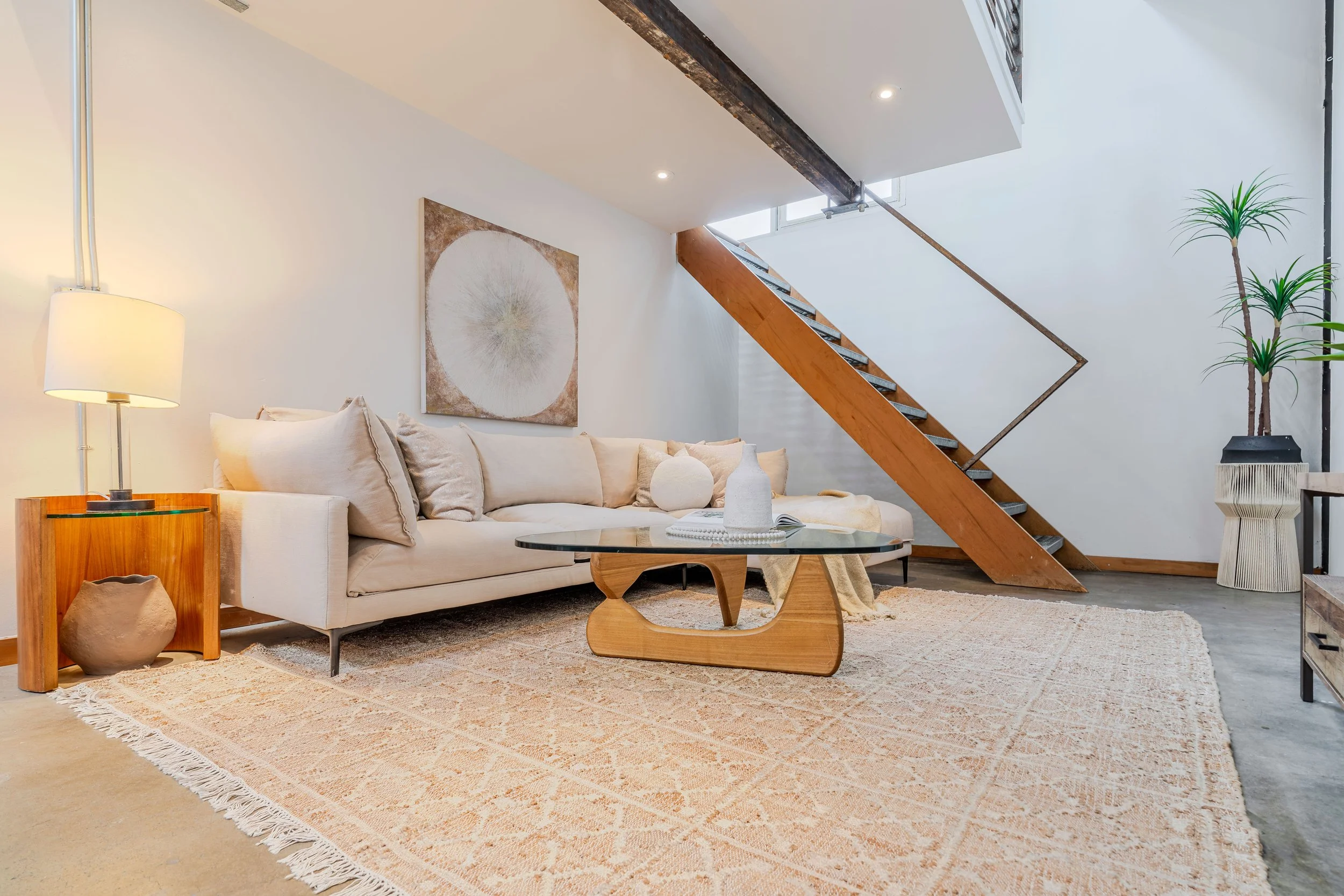 Living room with white sofa, glass coffee table, abstract artwork on the wall, wooden staircase, tall indoor plant, side table with lamp, and a beige rug.