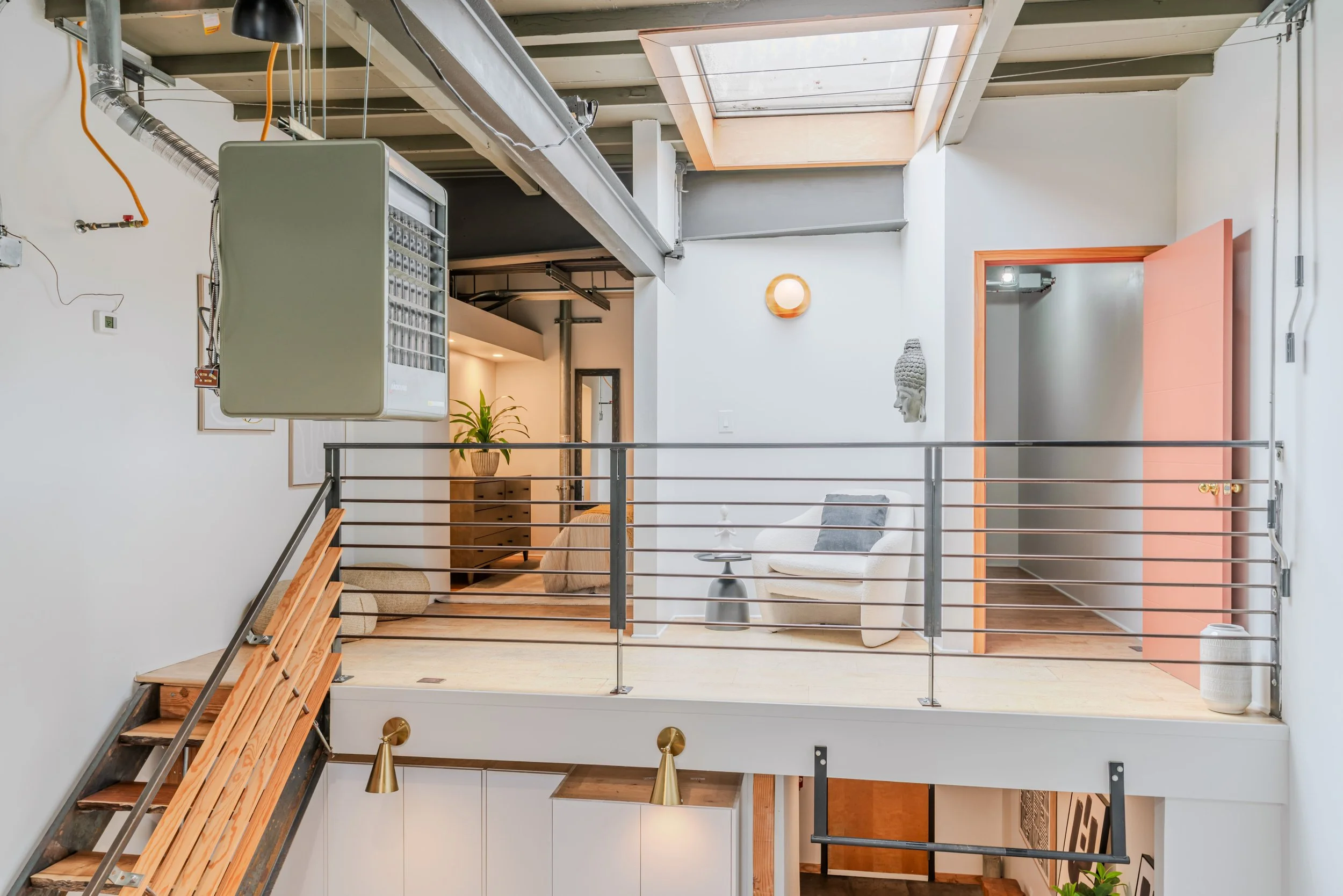 Interior view of a modern, open-concept living space with a mezzanine, featuring a sitting area with a beige sofa, a side table, wall art, and decorative plants, with a staircase in the foreground.