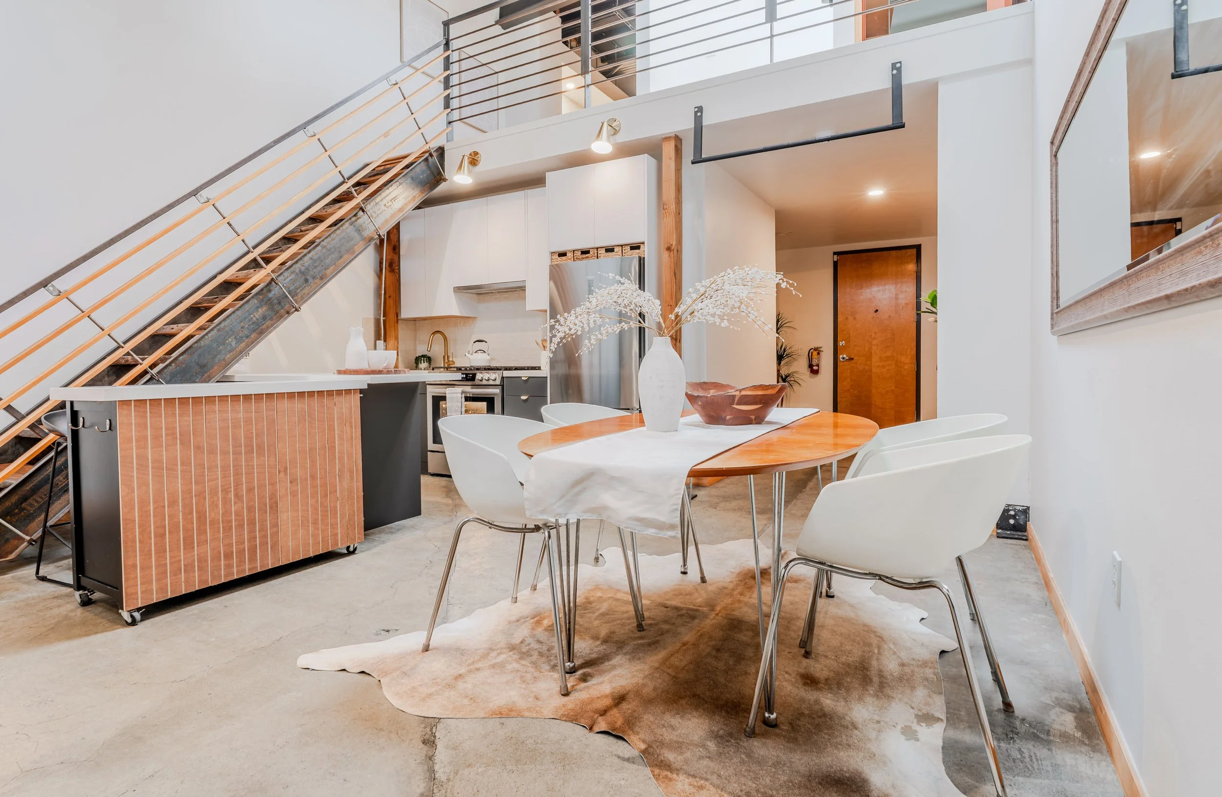 Interior of a modern apartment with an open kitchen and dining area. There is a round wooden table with white chairs, a white vase with flowers, and a cowhide rug on the concrete floor. The kitchen has white cabinetry, stainless steel appliances, and