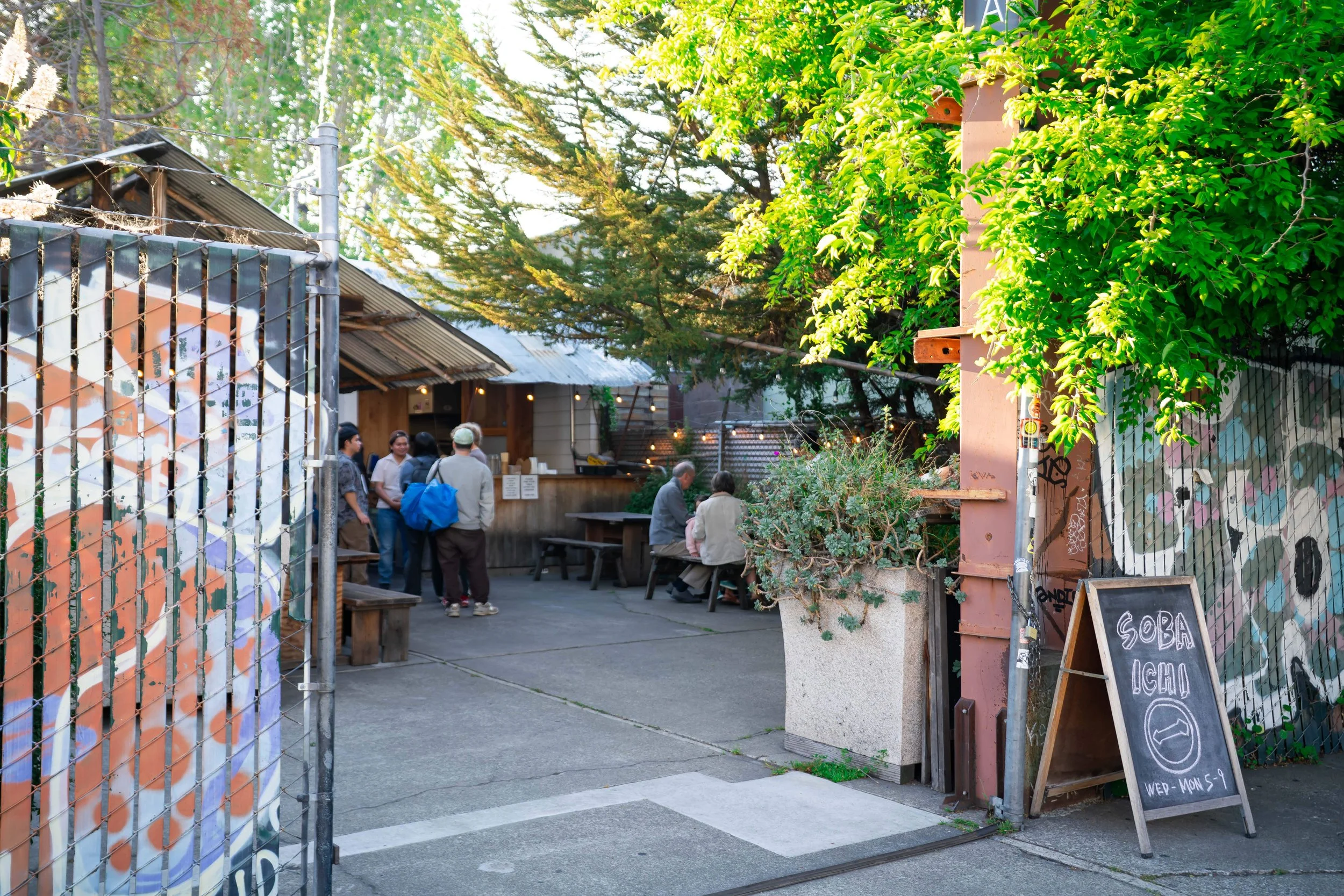 An outdoor scene at a cafe or restaurant entrance with a chain-link fence, a chalkboard sign, and a bamboo divider, with people gathered near the counter and sitting at tables under trees.