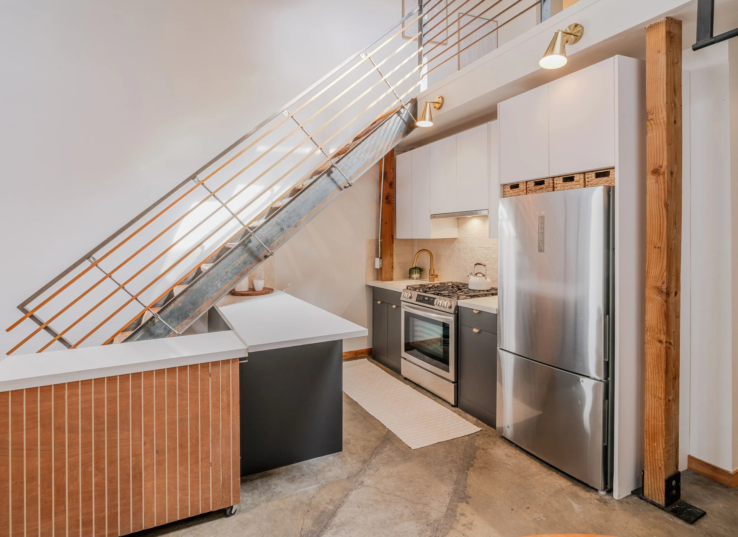Modern kitchen with white walls, wooden accents, stainless steel refrigerator, stove, and decorative lighting. A metal staircase with wooden slats is visible.