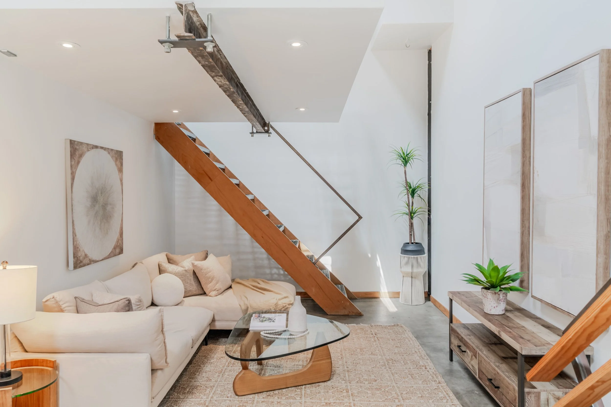 Living room with white walls, beige sofa, round glass coffee table, potted plants, wall art, wooden stairs, and contemporary decor.