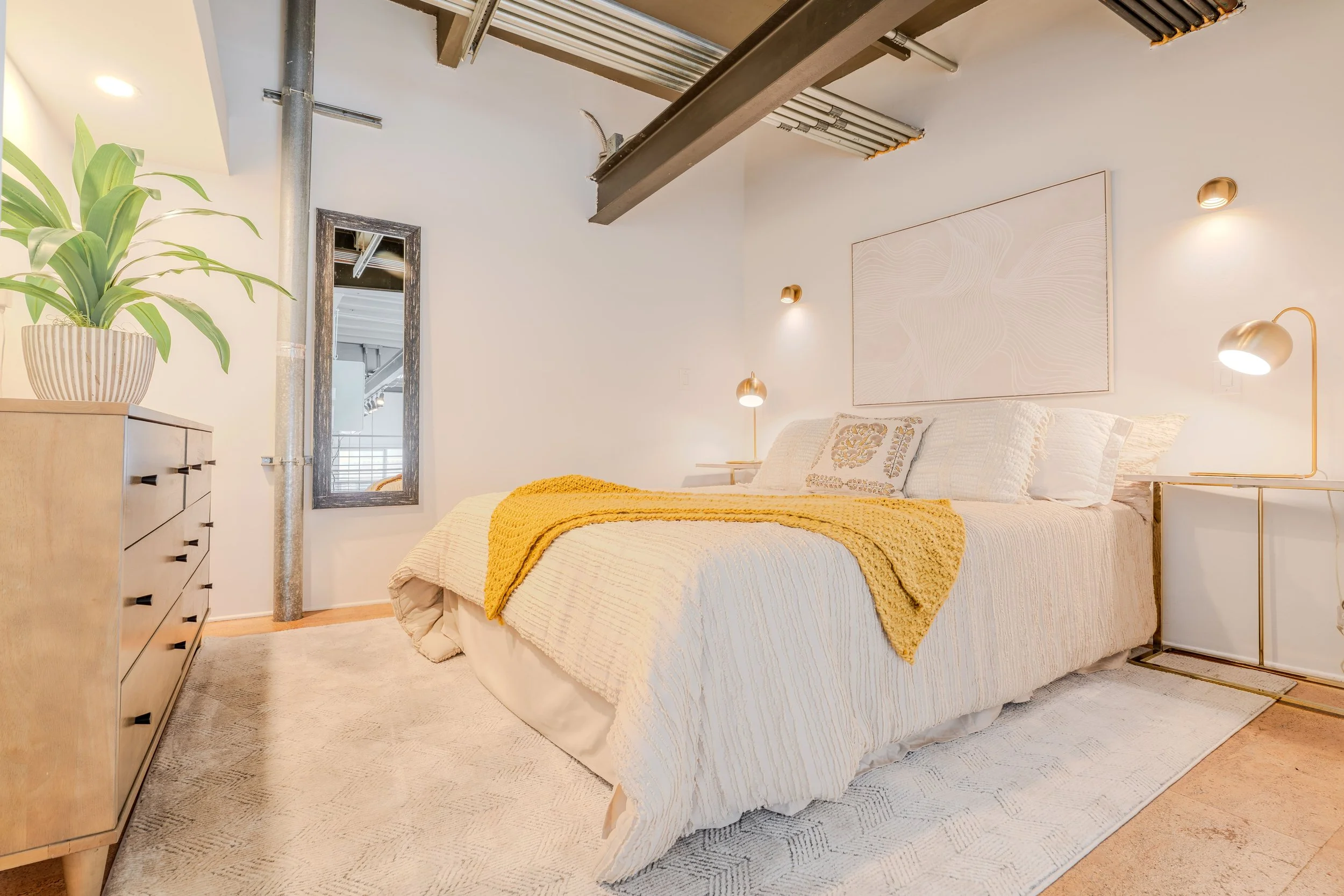Modern bedroom with a white bed dressed in white bedding and a yellow throw blanket. A wooden dresser with a potted plant is nearby. Wall lamps and a large abstract artwork in soft tones decorate the white wall. A mirror and exposed ceiling pipes add