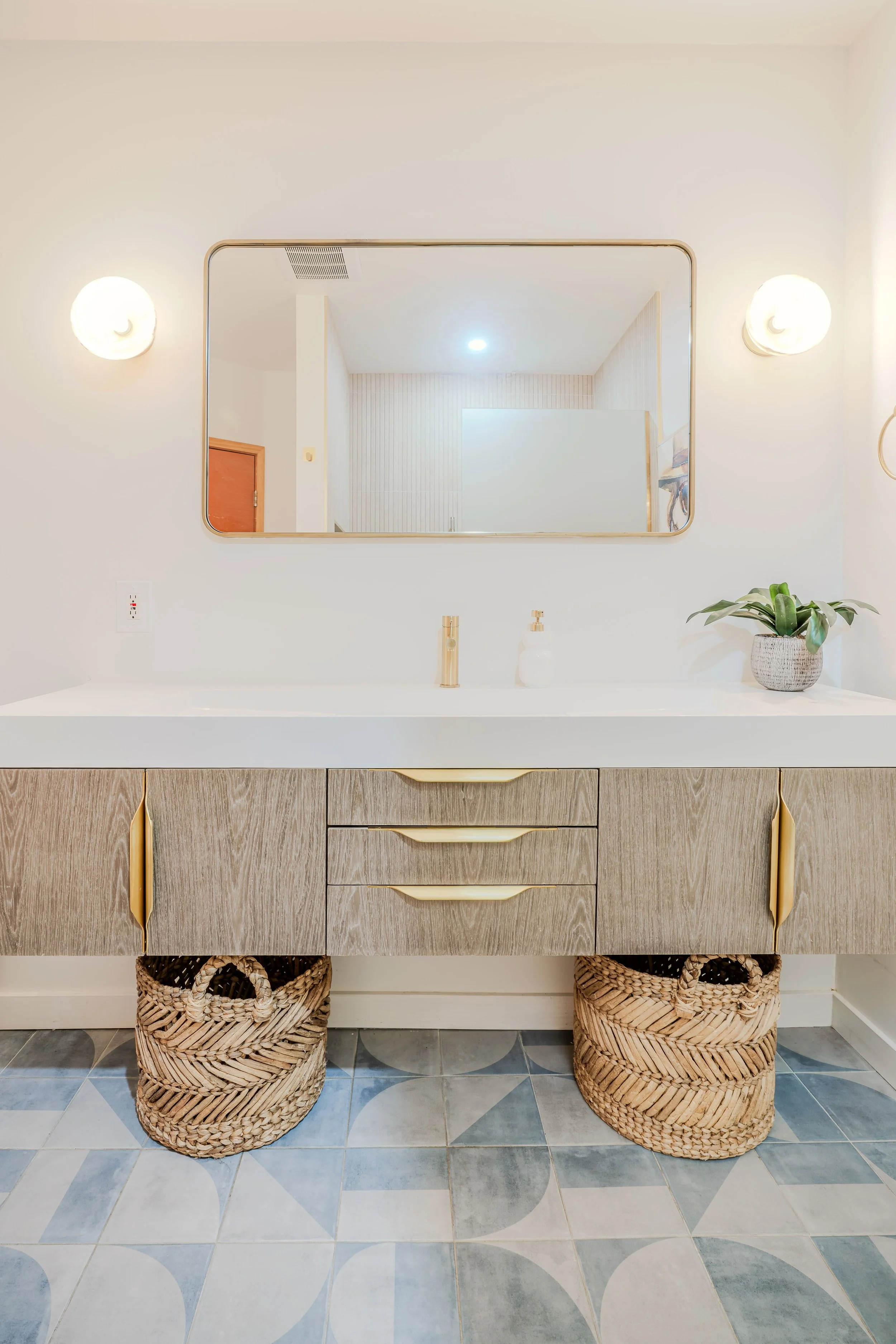 Bathroom vanity with a light-colored wooden cabinet, a white countertop, and woven baskets underneath. A large rectangular mirror is mounted above the sink, flanked by two wall lights. A small potted plant and soap dispenser are on the countertop. Th