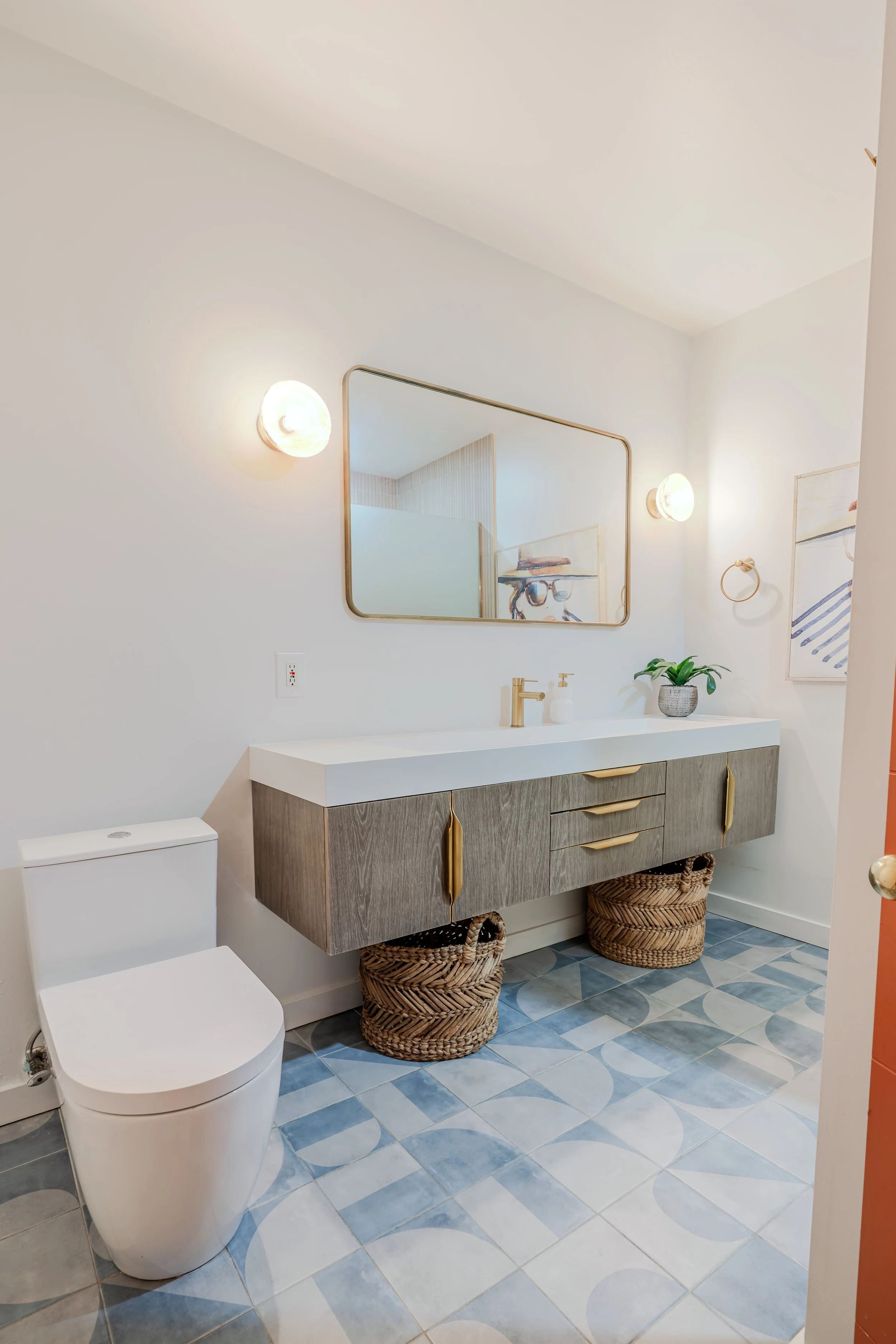 Modern bathroom with a white toilet, a floating wooden vanity with a white countertop and a gold faucet, a plant in a gray pot, and two woven baskets underneath. Mirror with golden frame and wall-mounted lights on either side of it, with a piece of w
