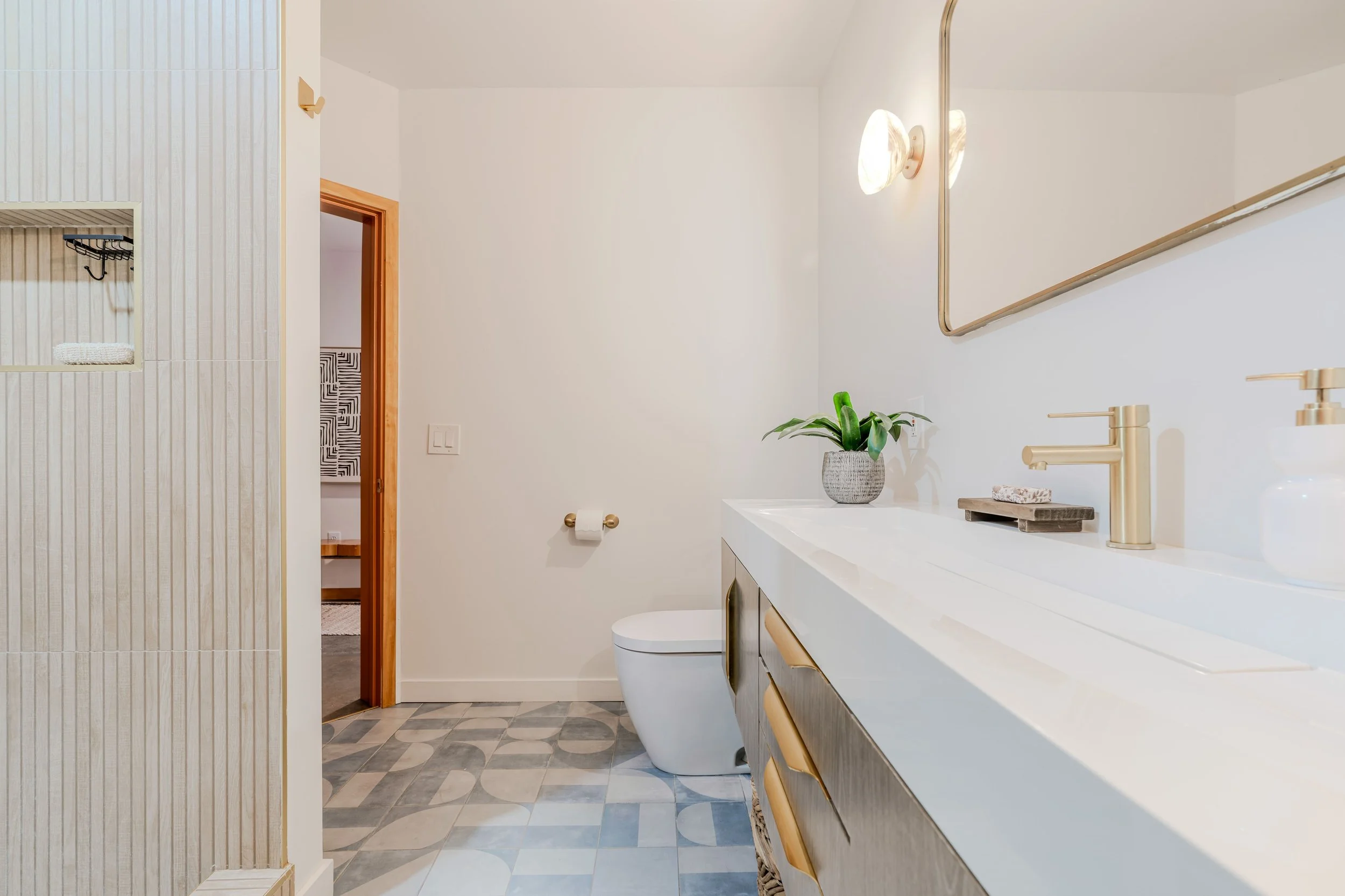 Modern bathroom with white walls, a vanity with gold accents, a potted plant, a gold faucet, wall-mounted light fixtures, a mirror, and a patterned tile floor.