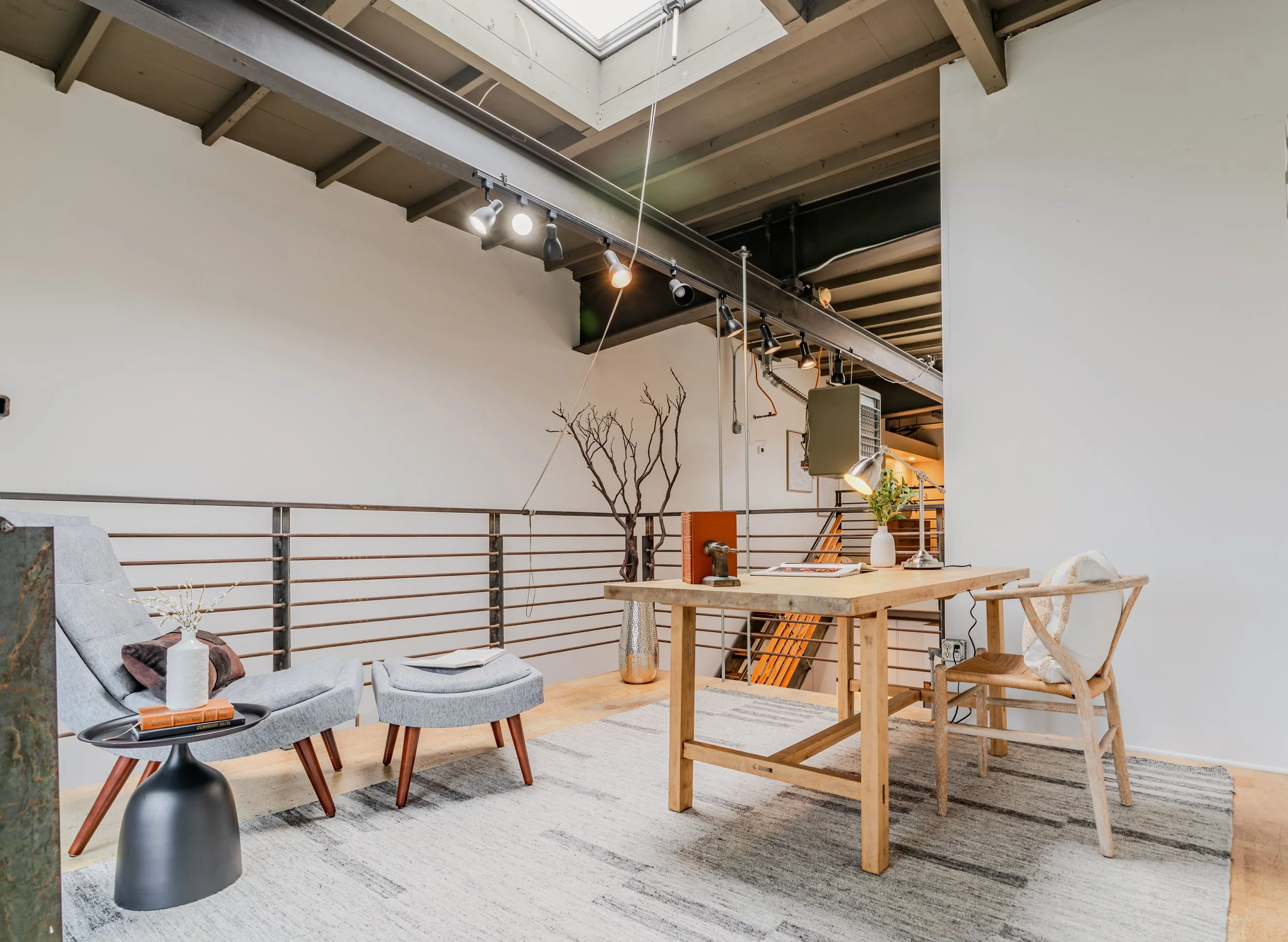 A modern loft-style living space with a high ceiling, exposed beams, track lighting, a small, round black side table with books and a bottle, a light gray sofa with cushions, a wooden dining table with chairs, a potted plant, and decorative branches 