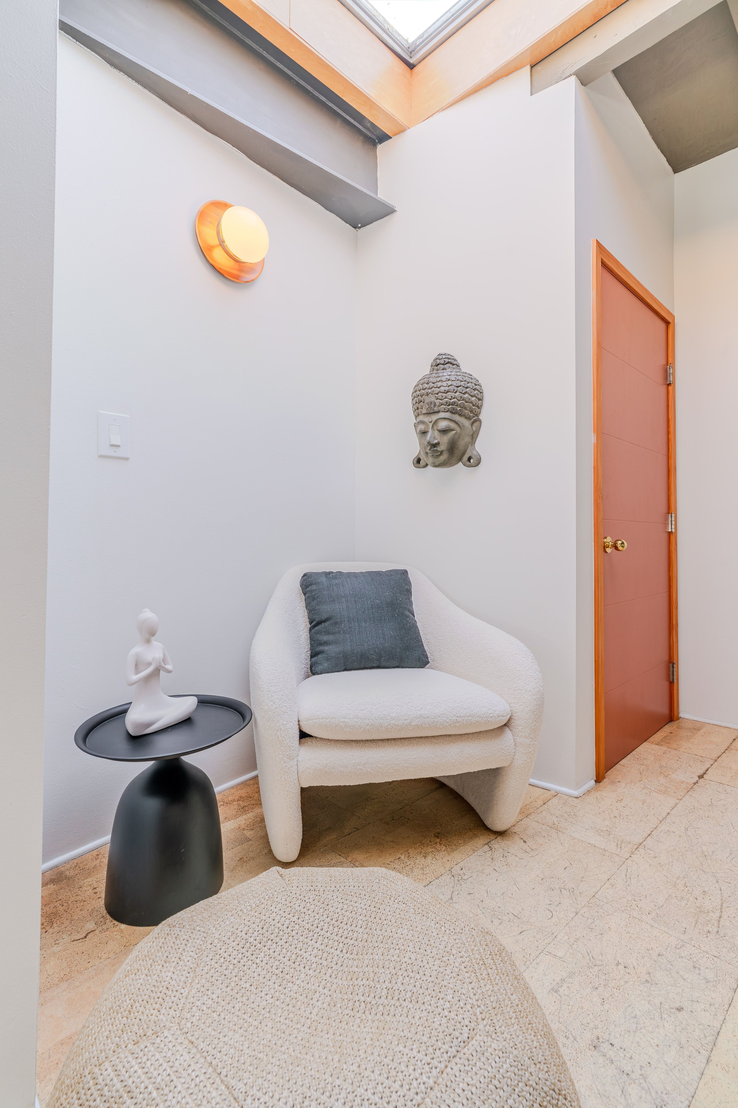A cozy corner with a white textured armchair that has a dark gray pillow, an oval side table with a white abstract sculpture of a woman, a wall hanging of a Buddha head, and a wall-mounted round light fixture, next to a pink door and a skylight.