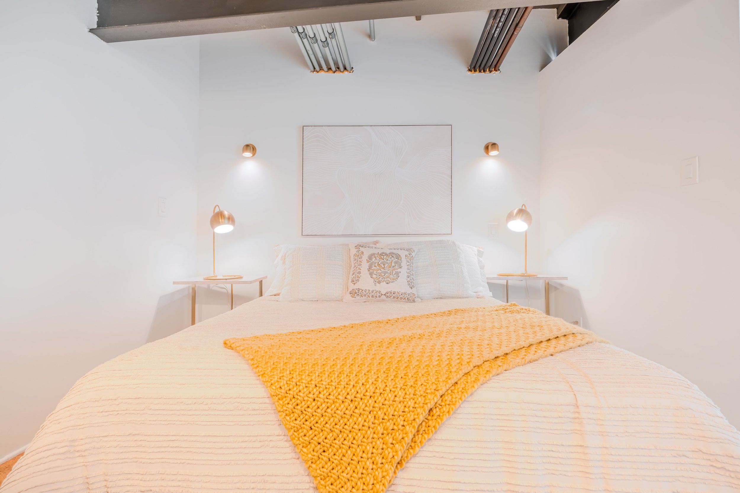 A neatly made bed with white bedding, a yellow knitted throw blanket, and decorative pillows, flanked by two bedside tables each with a copper-colored lamp. A minimalist abstract art piece hangs above the bed. The room features white walls and modern