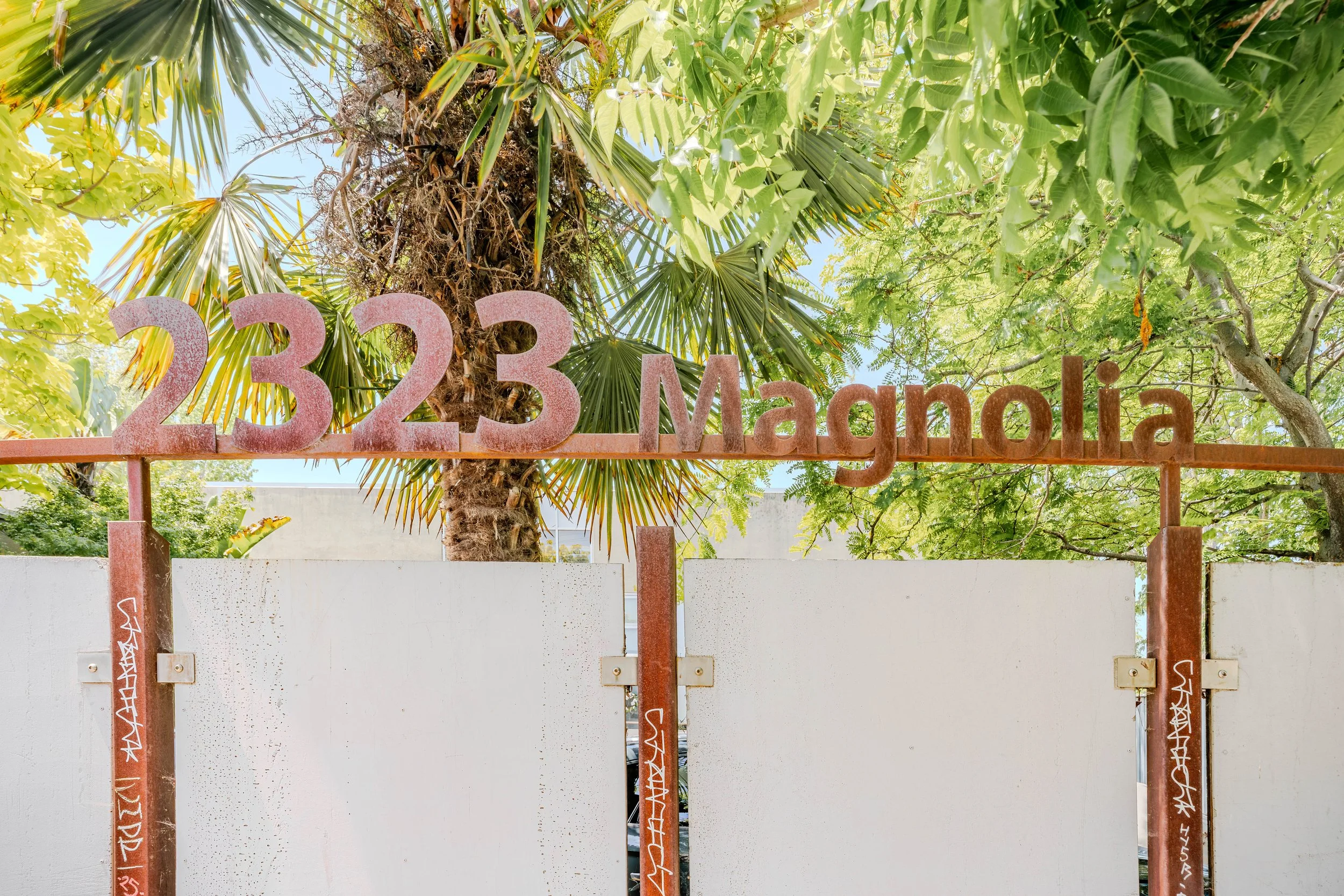 A decorative metal sign with the numbers and letters '2323 Margal' in pink, mounted on a metal framework, with a lush green tree and blue sky in the background.