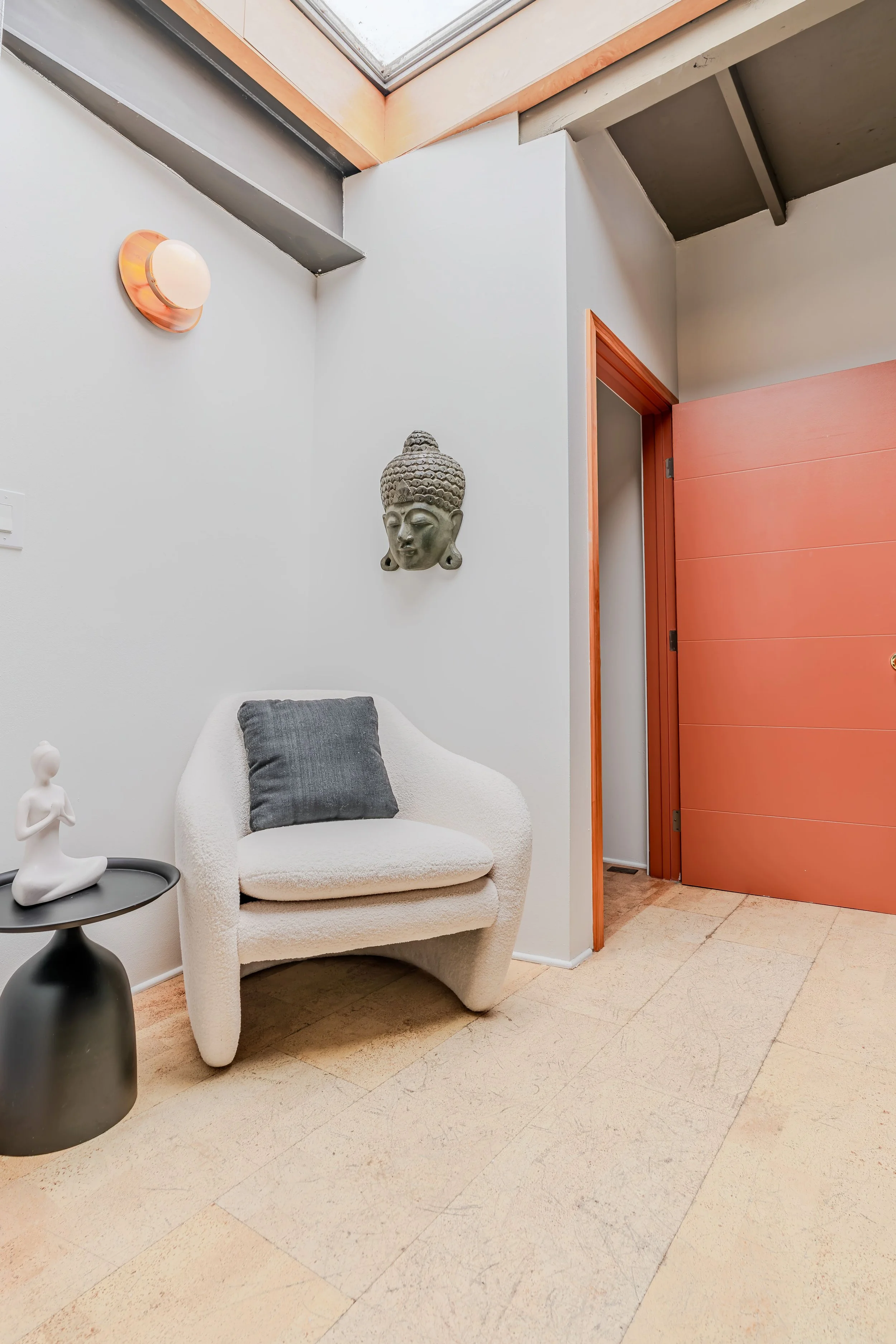 A cozy corner with a white plush armchair with a dark gray pillow, next to a small black side table with a white decorative figurine, against a white wall—decorated with a Buddha mask and a round wall sconce. A large rectangular window in the ceiling