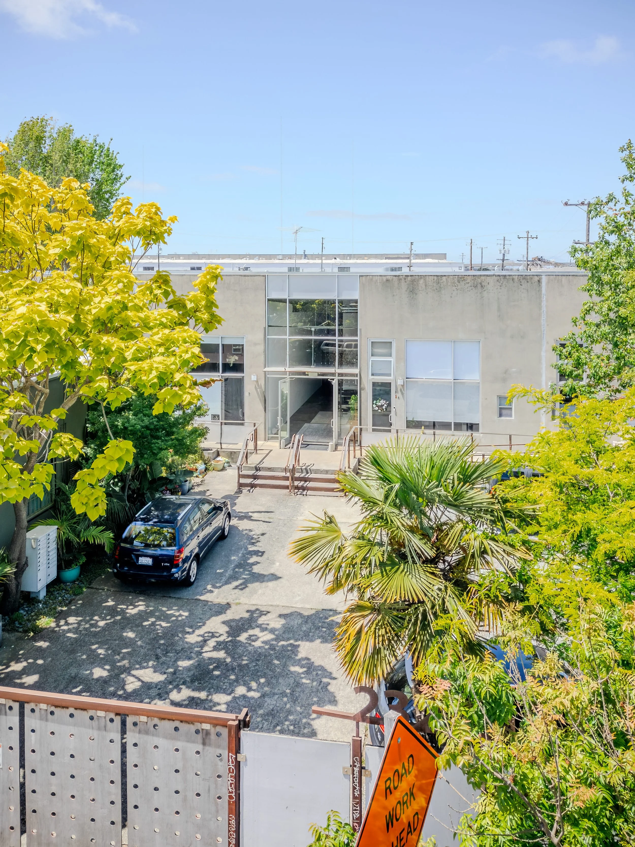 View of a building with glass doors and windows, surrounded by green trees, parked cars, and a street sign indicating road work ahead. The sky is blue with some clouds.
