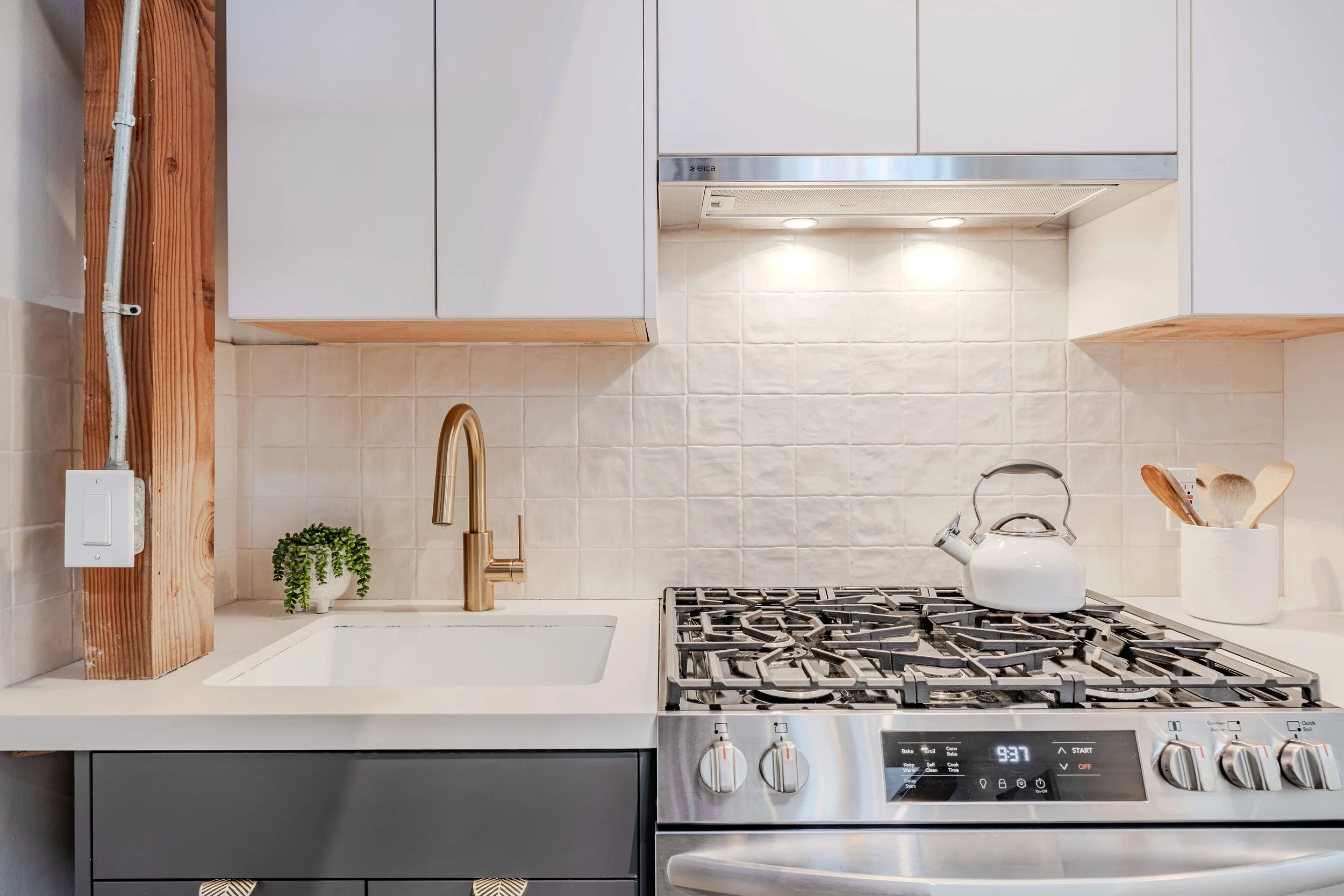 Modern kitchen with white cabinets, a brass faucet, a gas stove with a white kettle, a plant, and utensils in a white container, with beige tiled backsplash and a black and gray lower cabinet.