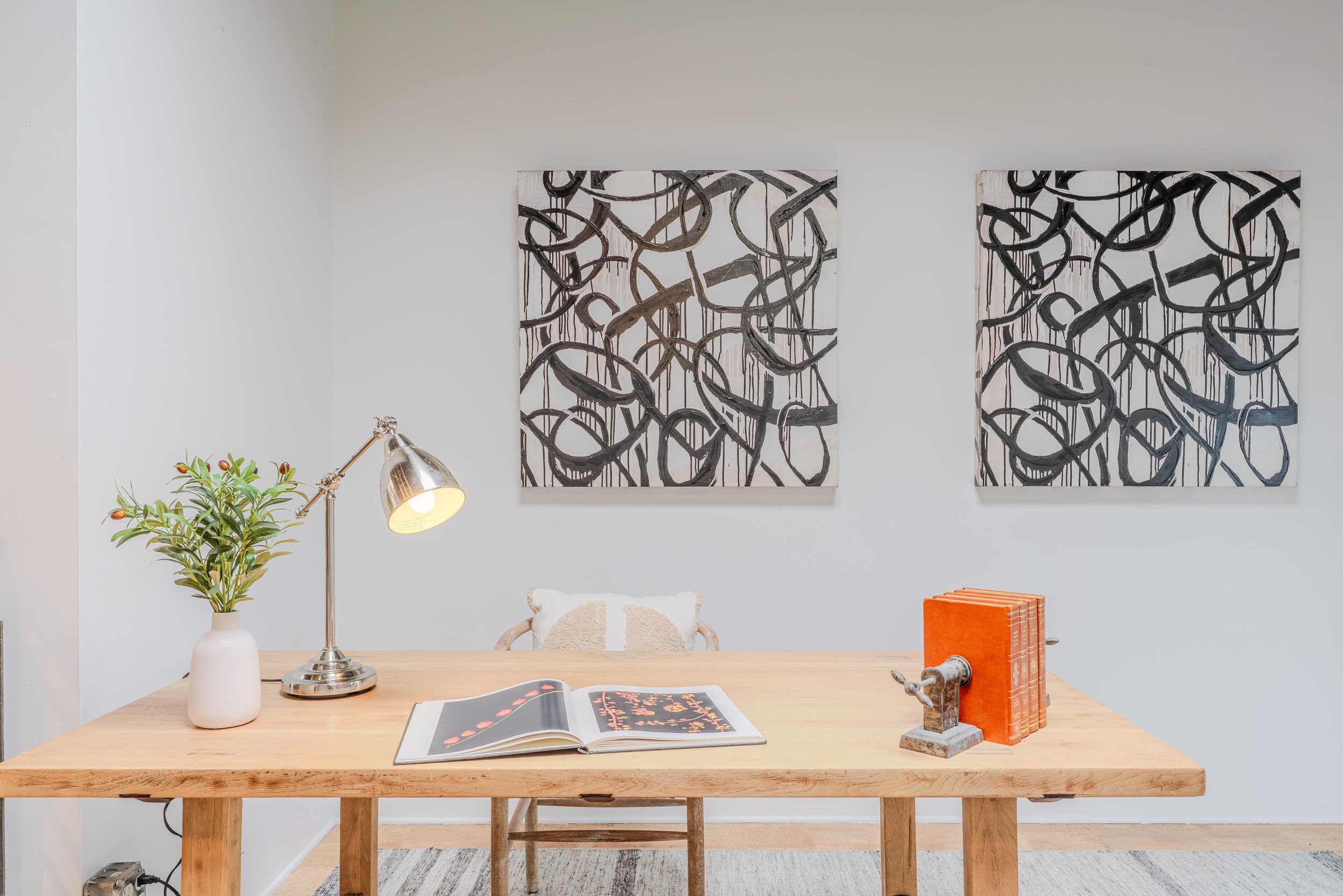 Interior of a modern office or living space with a wooden desk, a white vase with green plant, a silver desk lamp, an open photo book, orange books, and small decorative sculptures. Two abstract black and white paintings with looping patterns are on 