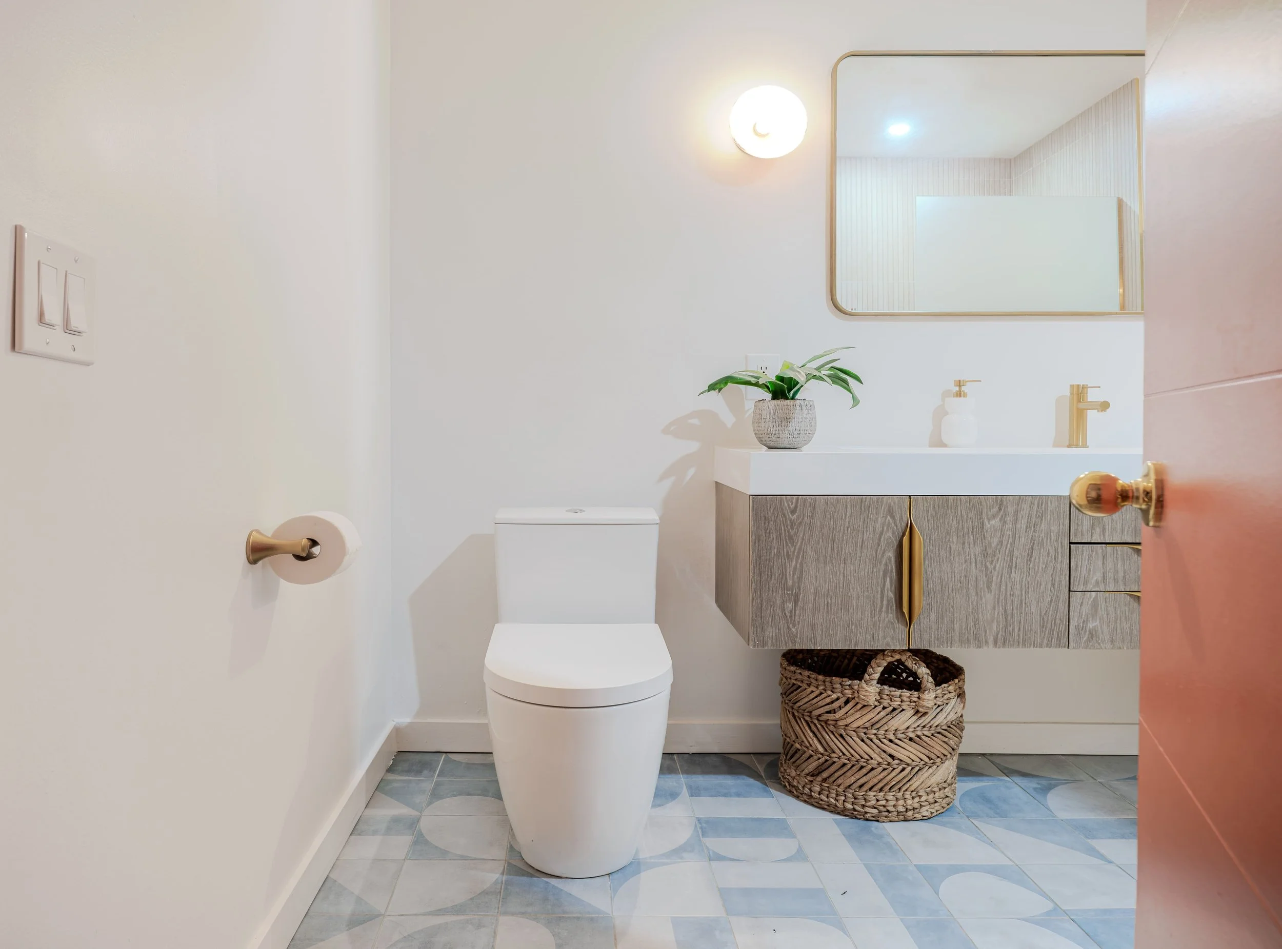 A modern bathroom with white walls, a round mirror, a small vanity with a gold faucet, a potted plant, a woven basket, and a toilet.