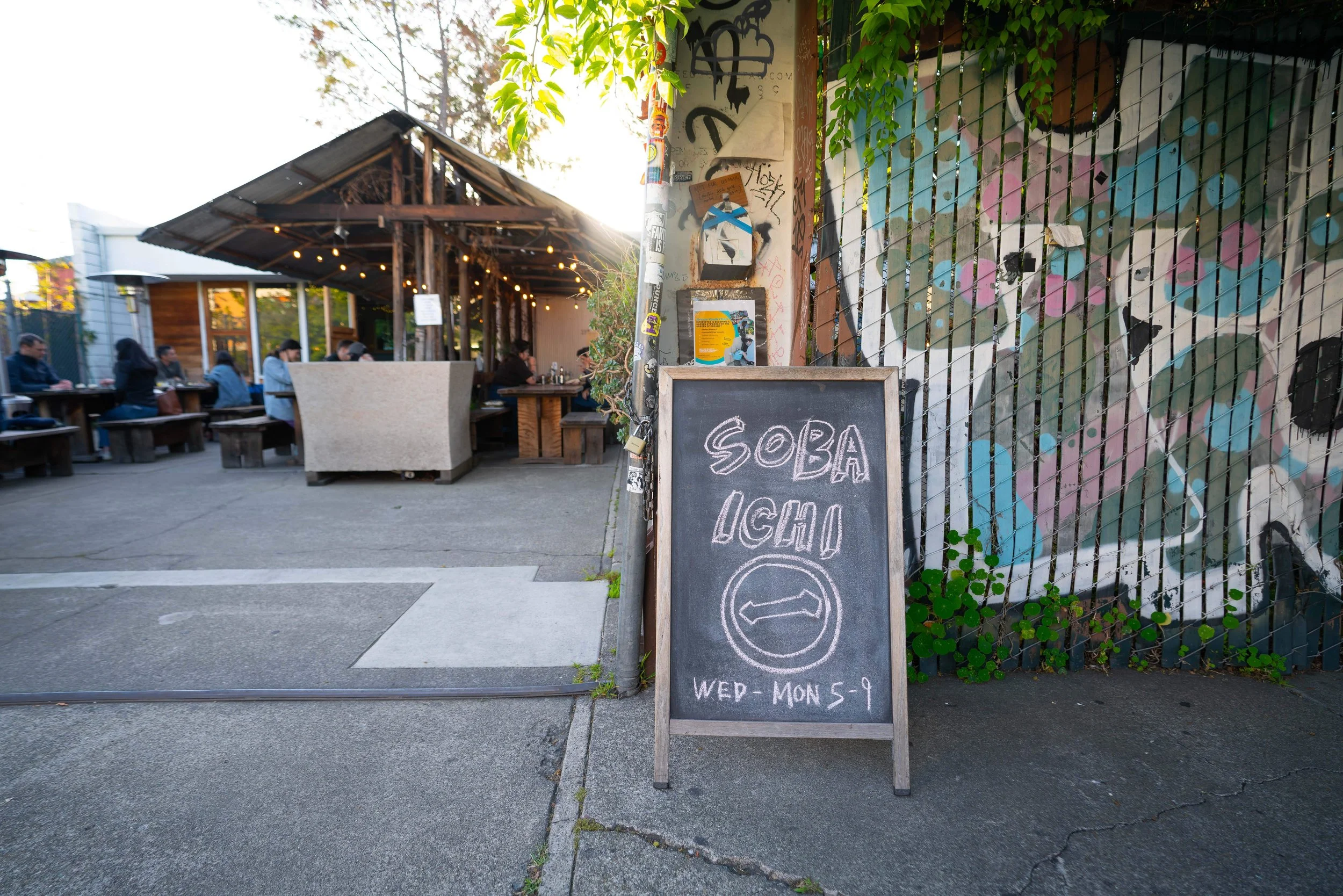 Outdoor restaurant patio with wooden tables, people sitting and dining under a black metal roof, surrounded by graffiti-covered fence, chalkboard sign advertising soy-based ice cream, and string lights, with sunlight and trees in the background.