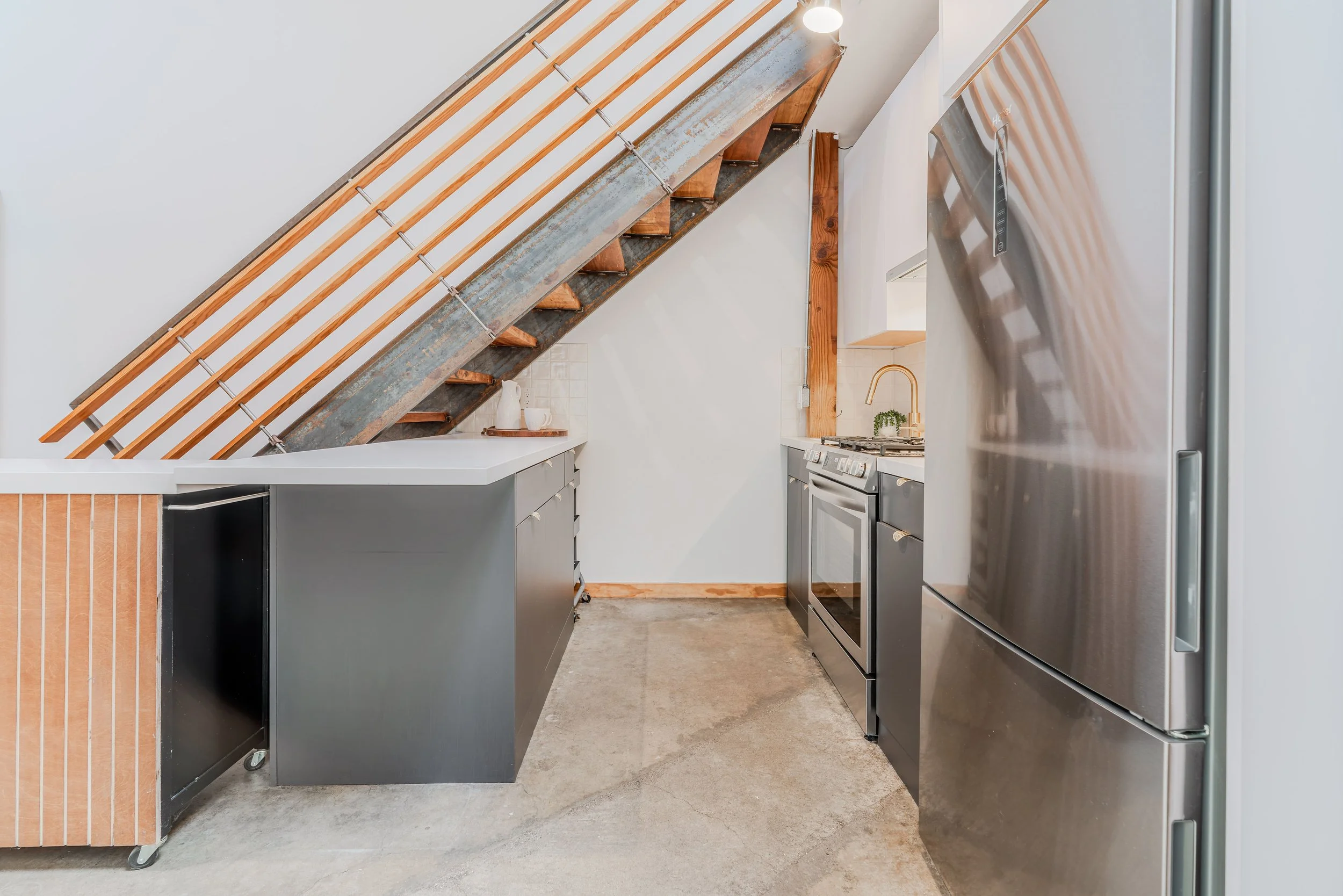 A modern kitchen with exposed staircase under the stairs, grey cabinets, stainless steel refrigerator, and concrete floor.