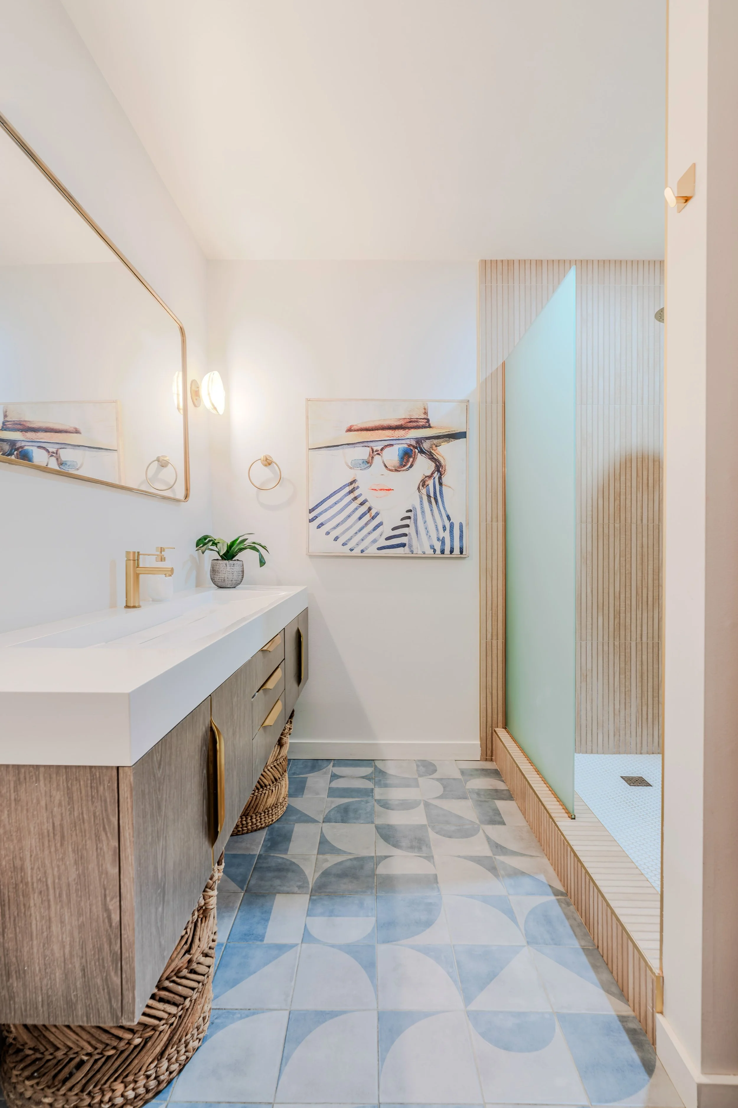 Modern bathroom with wooden vanity, woven basket, framed artwork of a woman with sunglasses, potted plant, wall lights, and a shower area with wooden and frosted glass panels, patterned tile floor.