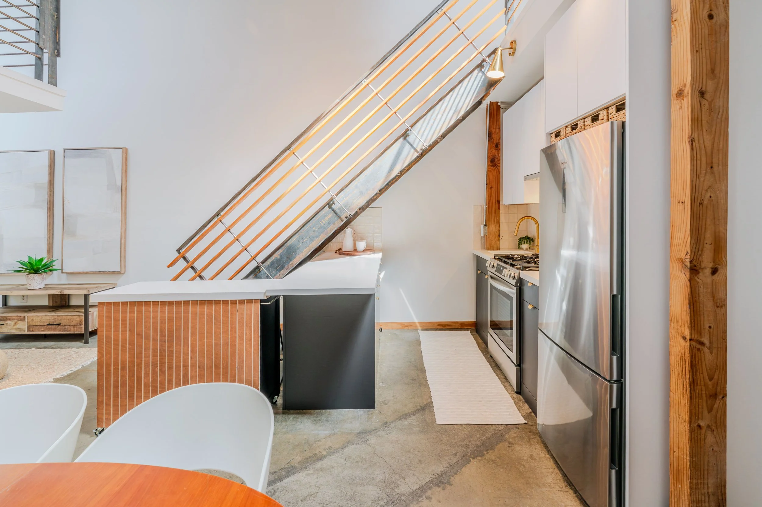 Interior view of a modern kitchen with stainless steel refrigerator, oven, modern sink with gold faucet, wooden accents, and a staircase with metal railing.