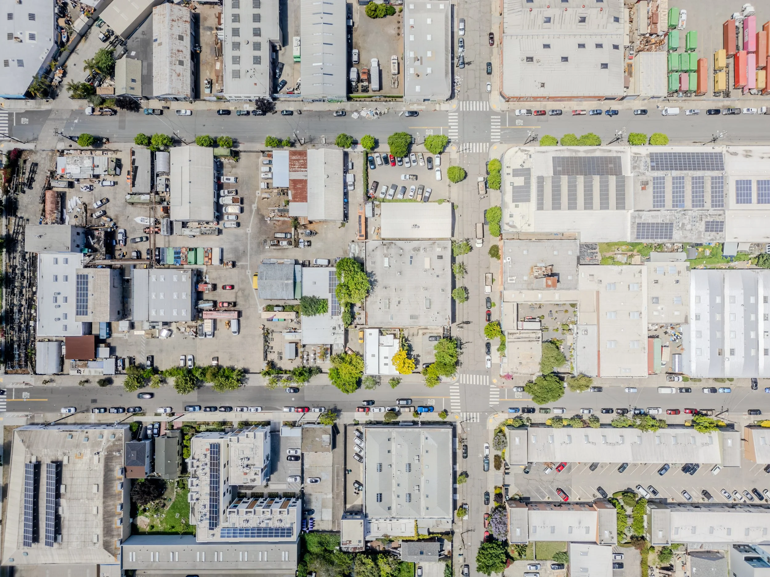 An aerial view of a parking lot and surrounding buildings on a city street, with cars parked and some trees lining the streets.