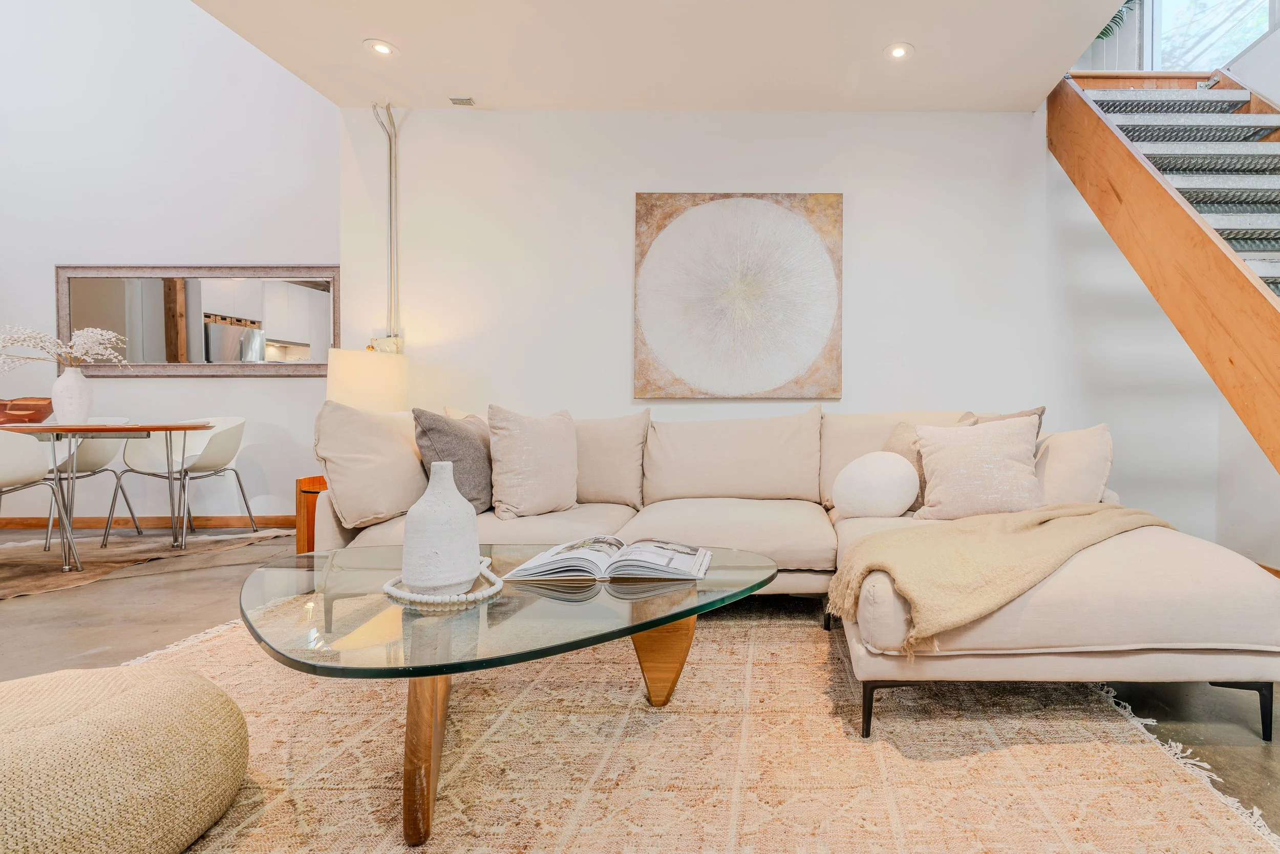 Living room with a beige sectional sofa, glass coffee table with a vase and an open book, walls with artwork, a lamp, and a staircase in the background.