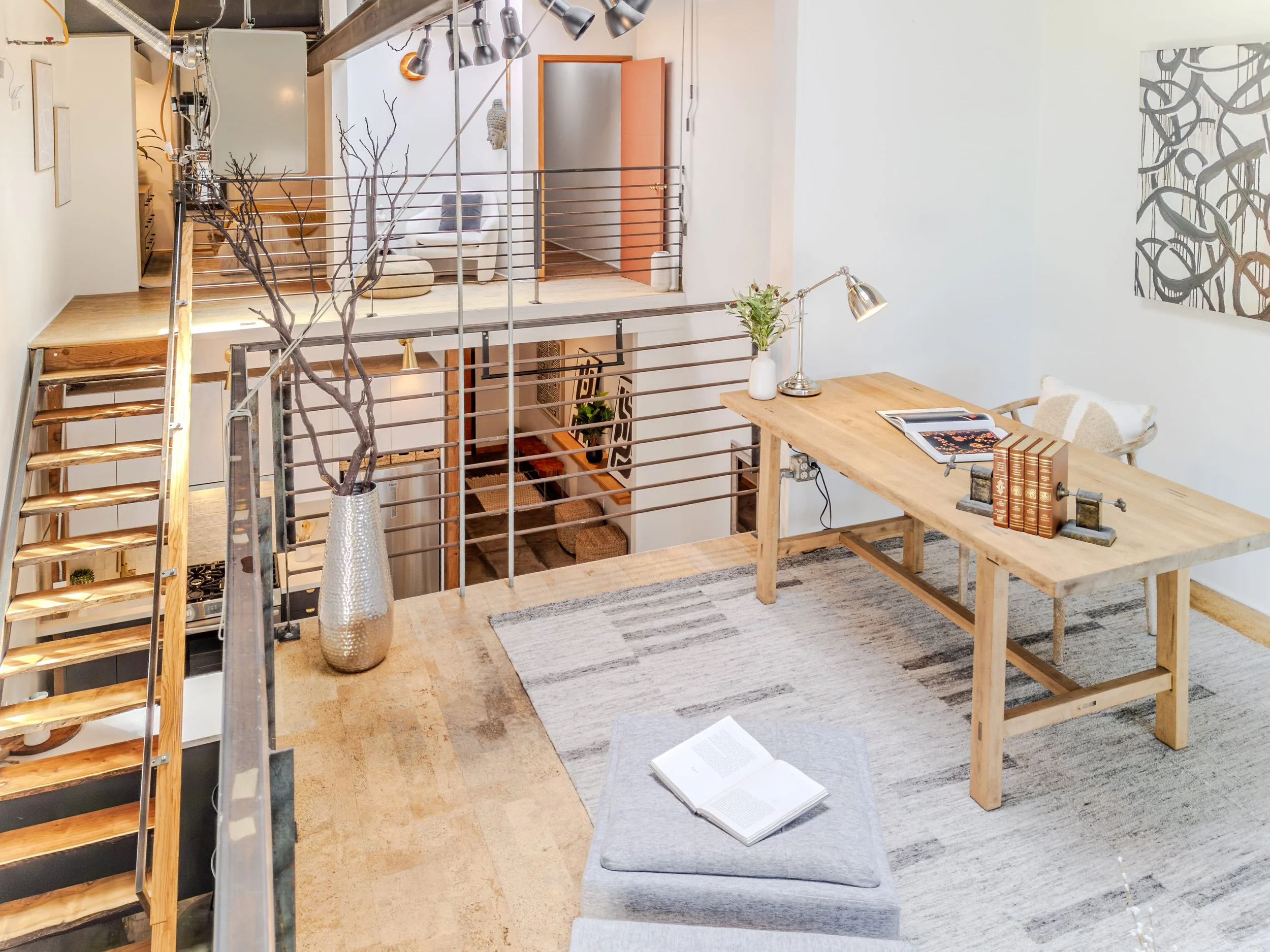 Interior of a modern home featuring a second-floor loft space with wooden floors and metal railing. There is a wooden table with a lamp, vase with flowers, and books. A staircase with wooden steps and metal railing leads downstairs. A large open book