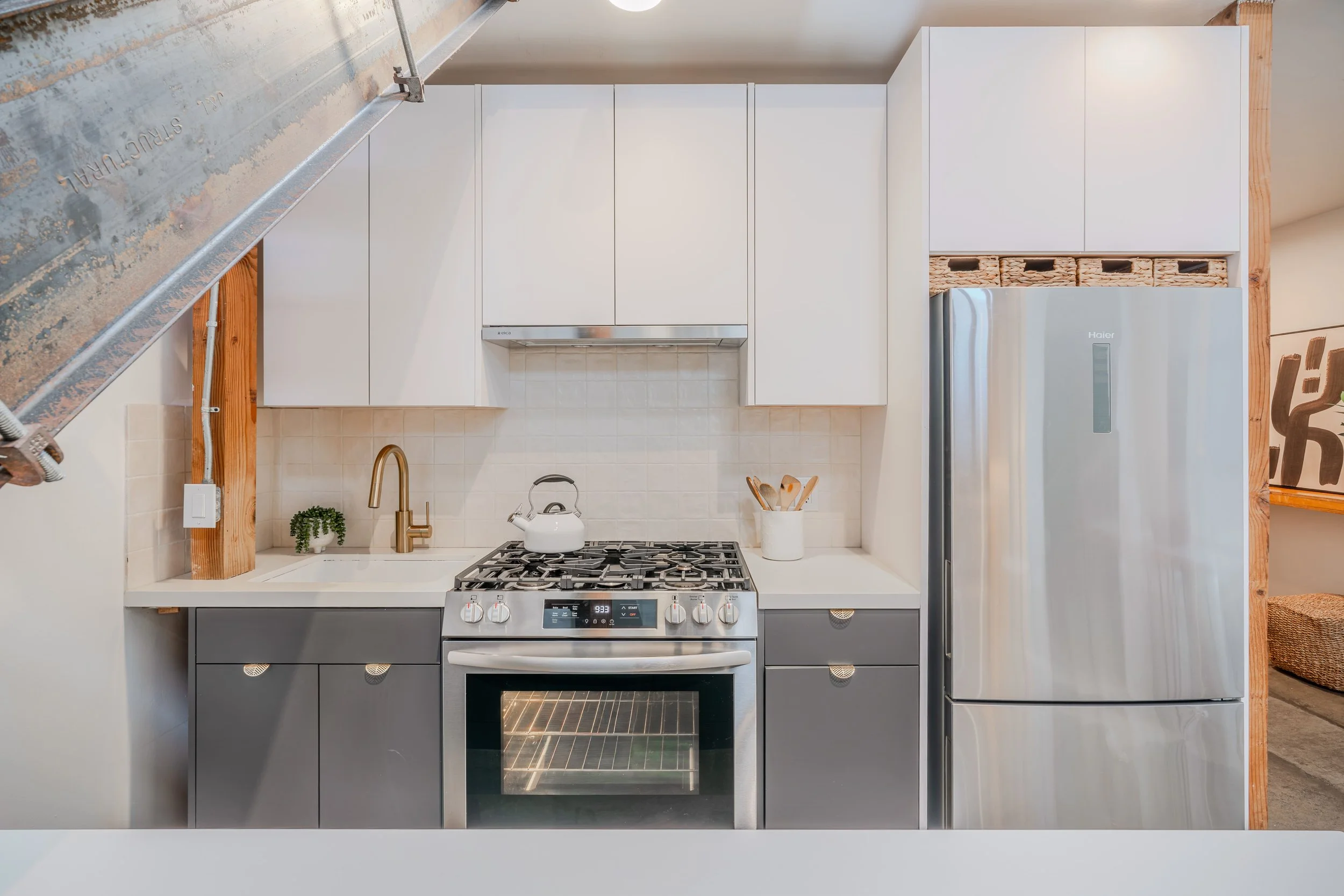 Modern kitchen with white cabinets, a stainless steel stove with oven, a white countertop with a kettle and utensils, a small plant, and a stainless steel refrigerator. Exposed wooden beams and light beige tiles on the backsplash.