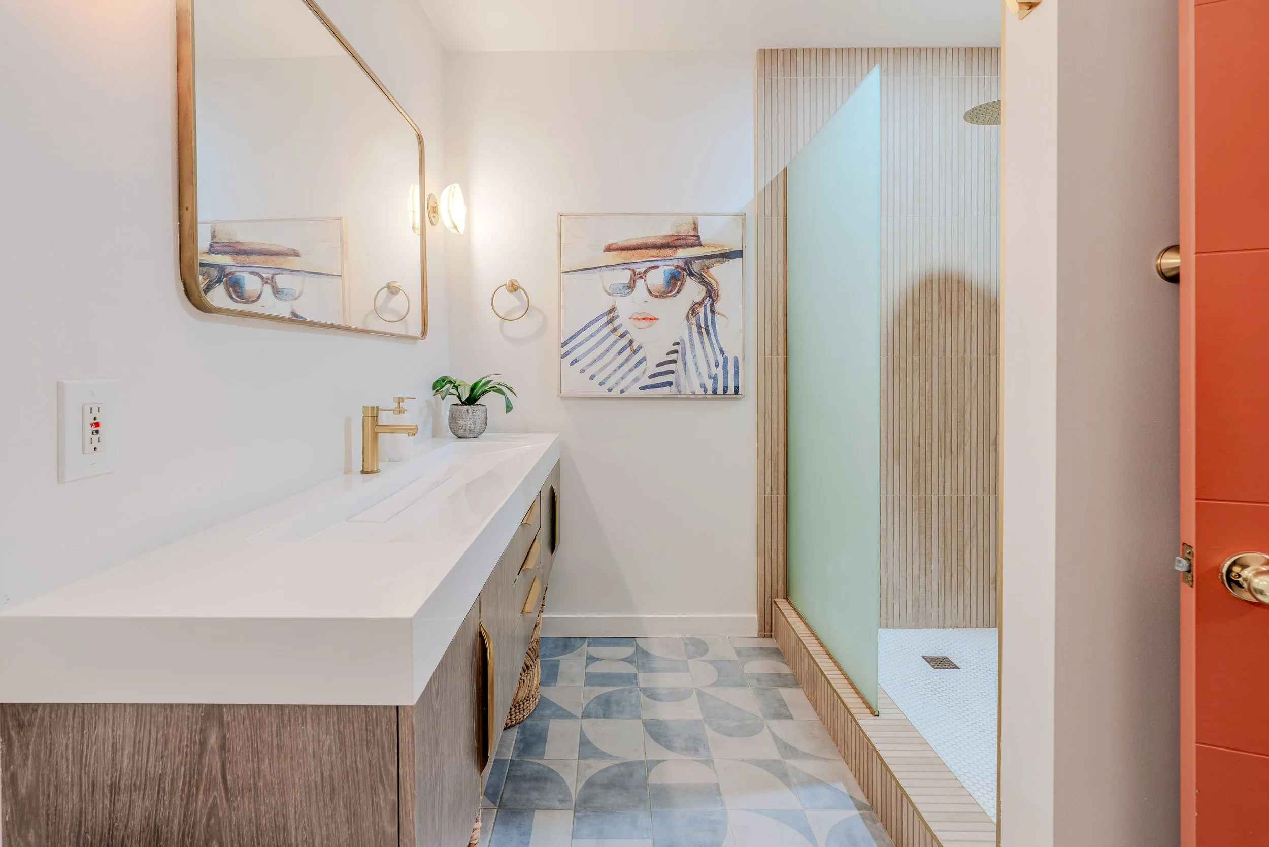 Modern bathroom with a white countertop, wooden cabinetry, a mirror, wall-mounted lights, and a shower area with a frosted glass partition, light gray patterned tile floor, and a partial view of an orange door.