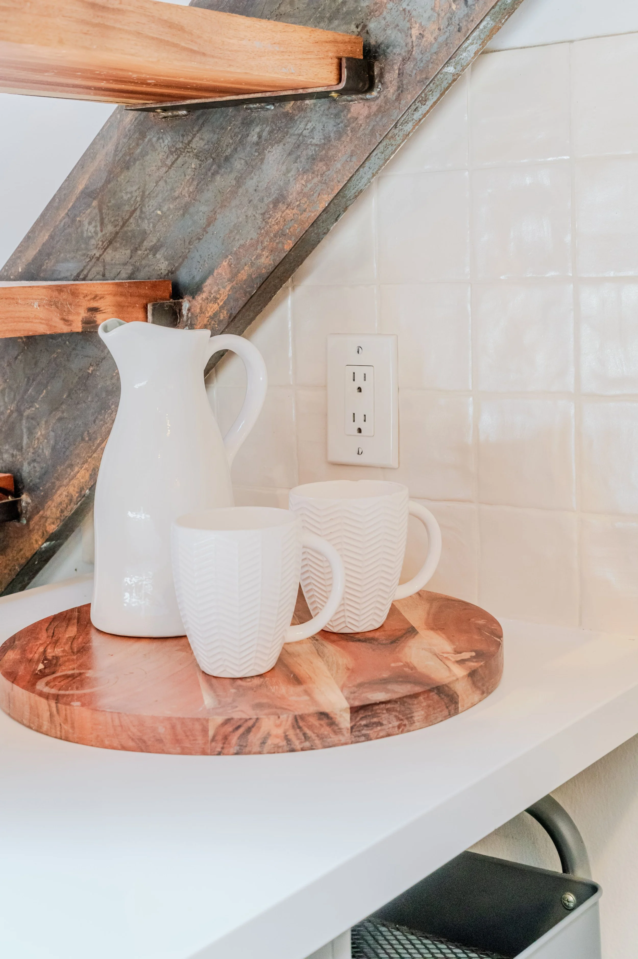 Three white ceramic mugs with textured patterns are placed on a round wooden tray, with a white ceramic pitcher behind them. A distressed wooden shelf and an electrical outlet are visible on the kitchen wall in the background.
