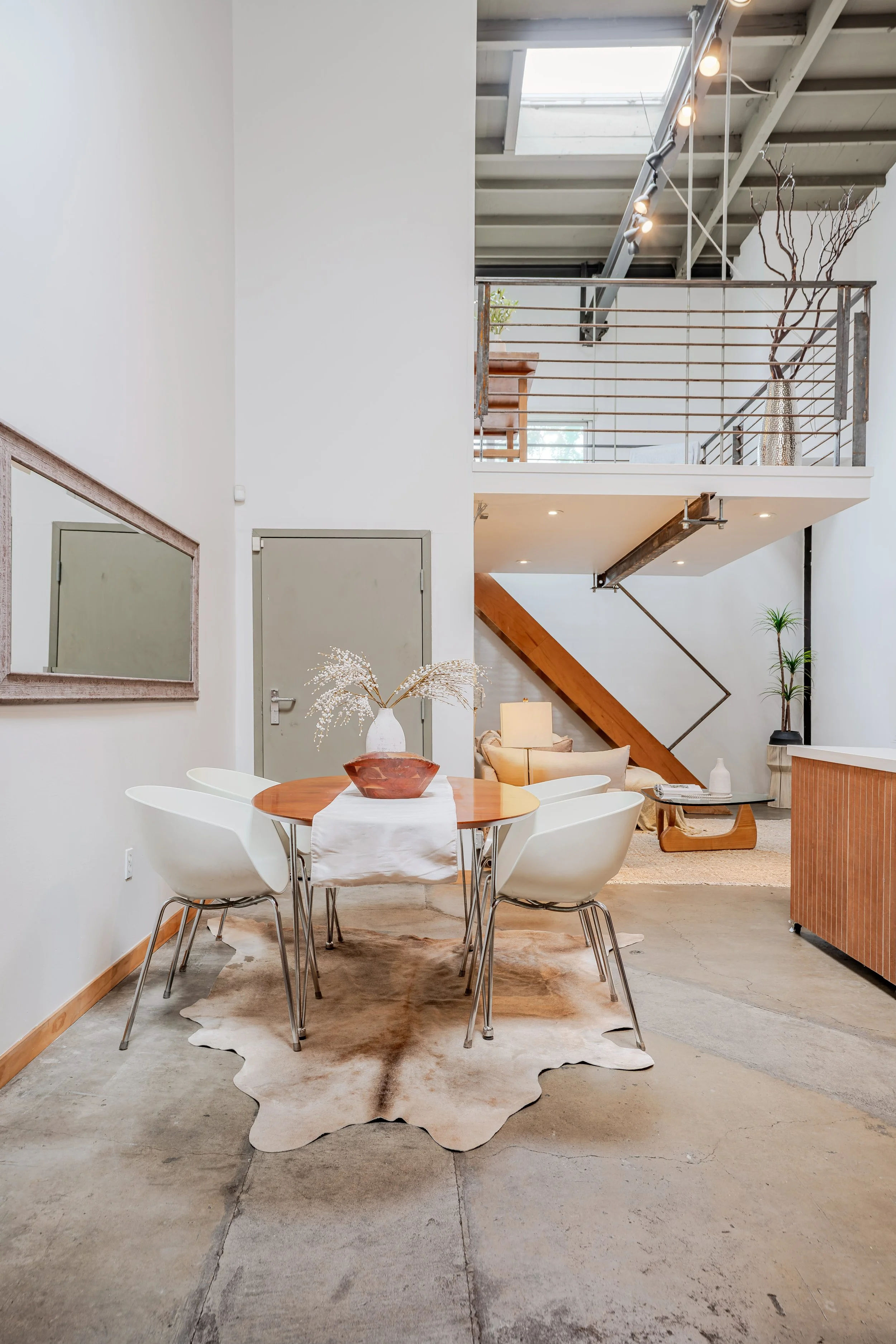 Modern interior of a home with a high ceiling, a staircase with wood steps and black metal railing, a dining area with a round wooden table and white chairs, a cowhide rug, and a gray door. The space has natural light coming from a skylight, and deco