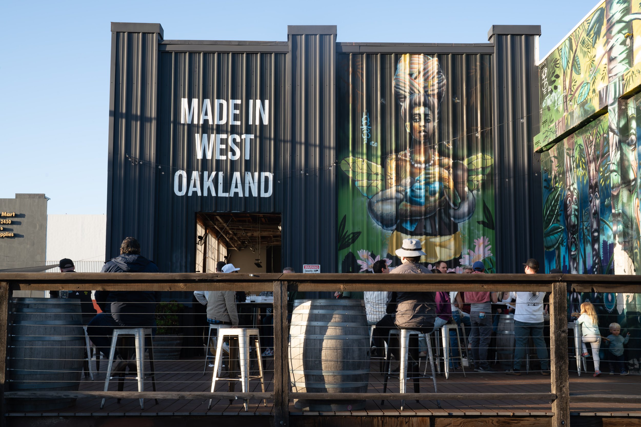 People gather outside a building with a mural of a woman with colorful hair and a serene expression, holding a blue object. The building has a sign that reads 'Made in West Oakland' and a partially open entrance. There are chairs, barrels, and a grou