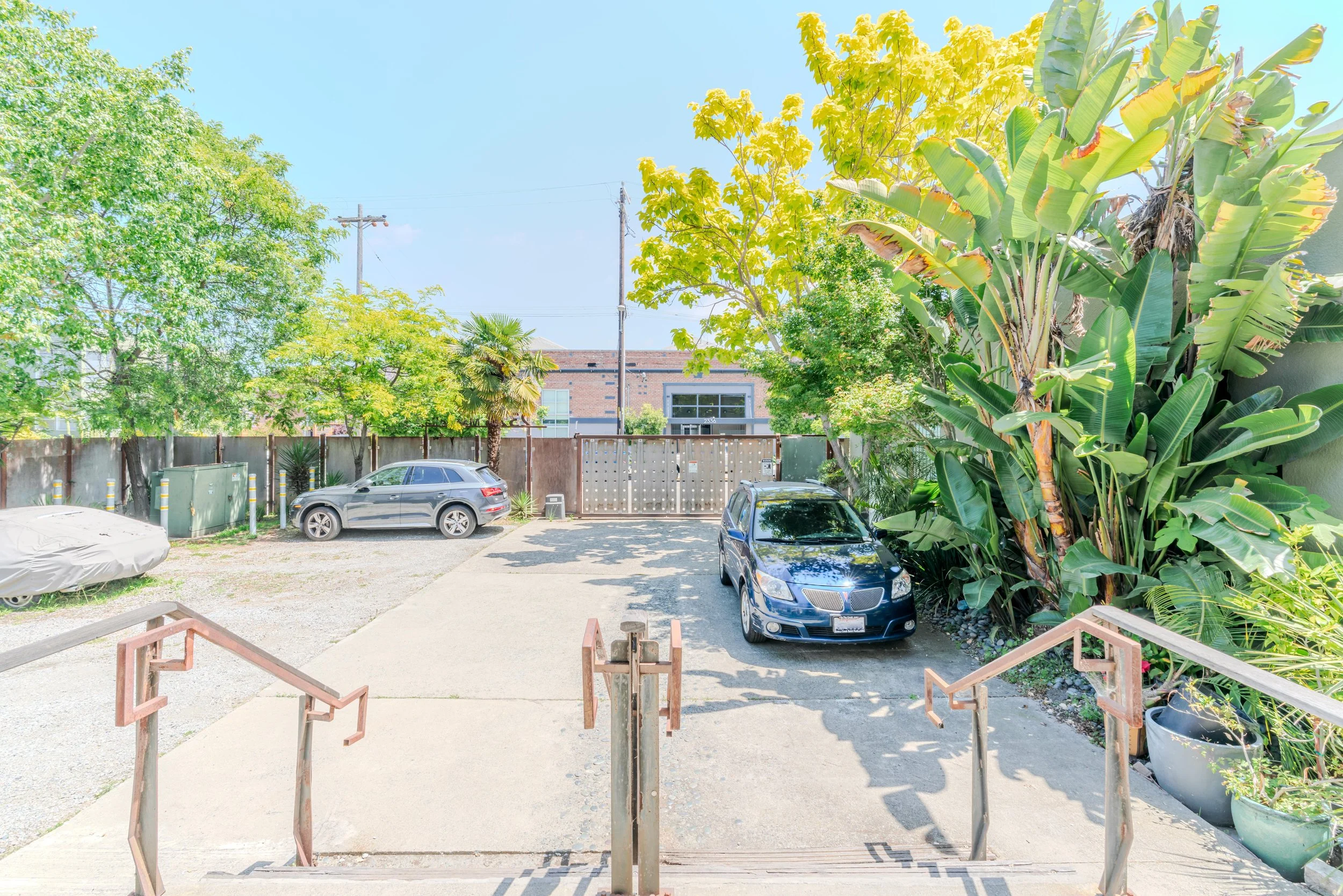 View of a parking area behind a building with three cars parked, one gray, one black, and one dark blue, surrounded by green trees and plants. A wooden fence and utility poles are visible in the background, with a brick building and a clear blue sky.