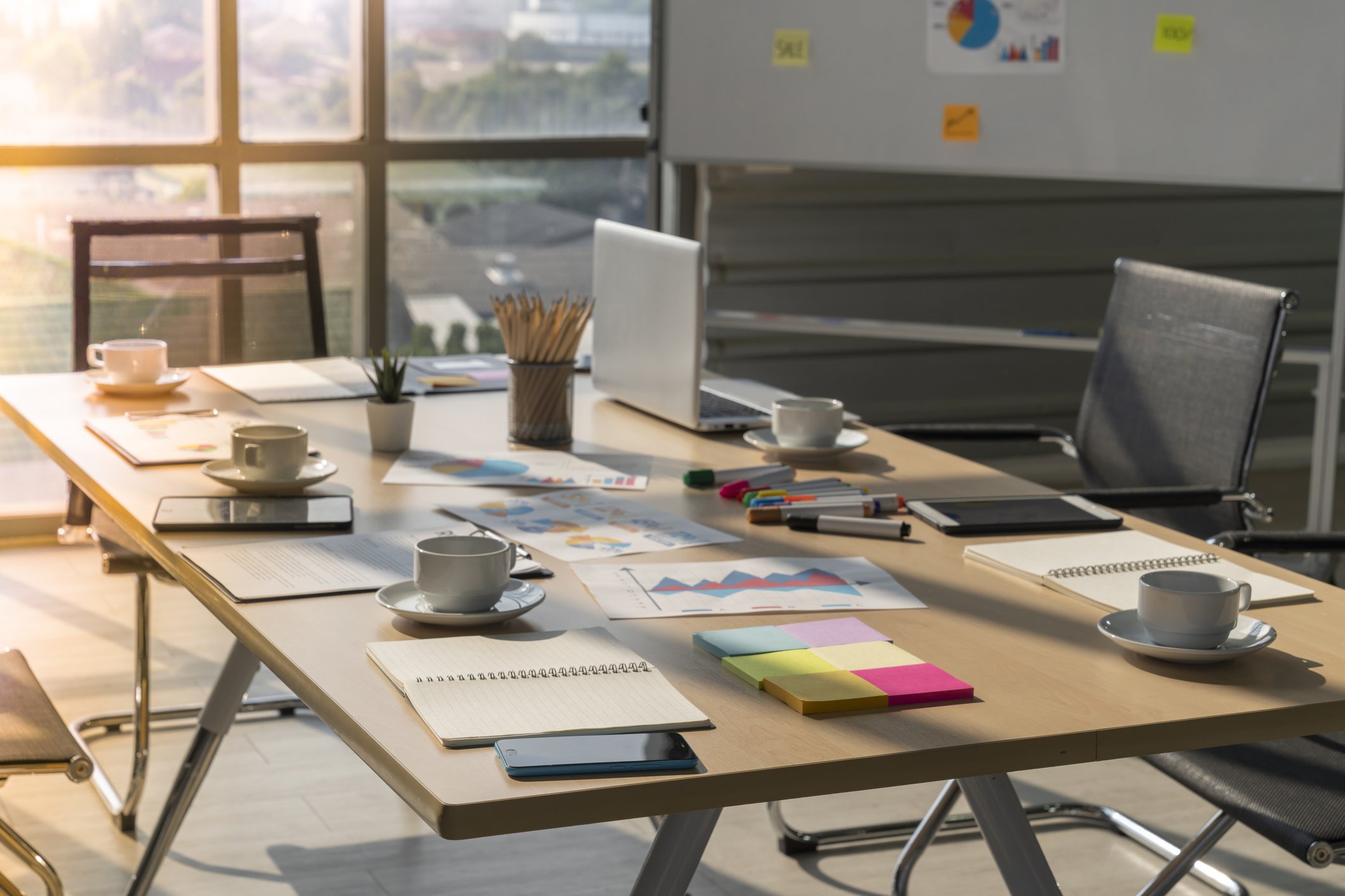 Office conference table with laptops, notebooks, coffee cups, colored markers, sticky notes, and documents, illuminated by sunlight through large windows.