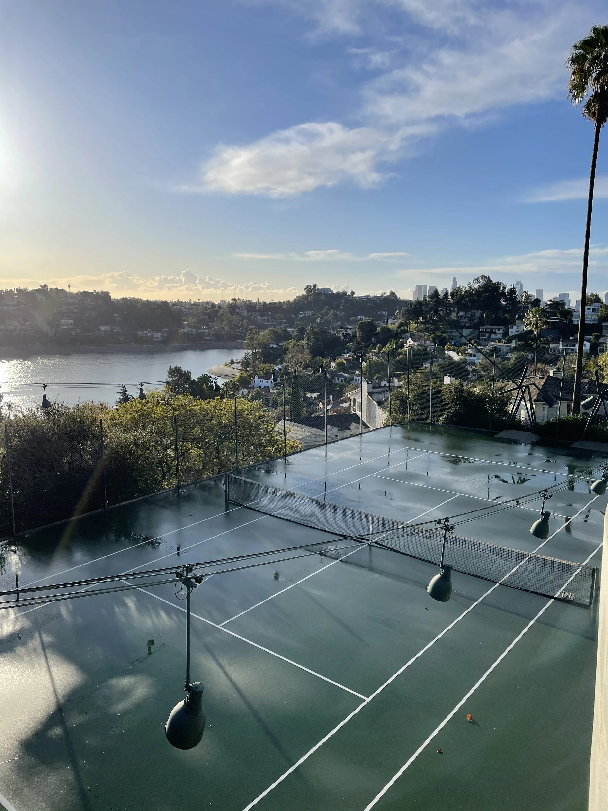 A tennis court with surrounding fences and lights, overlooking a body of water and a hillside with trees and houses, with a city skyline in the distance under a partly cloudy sky.
