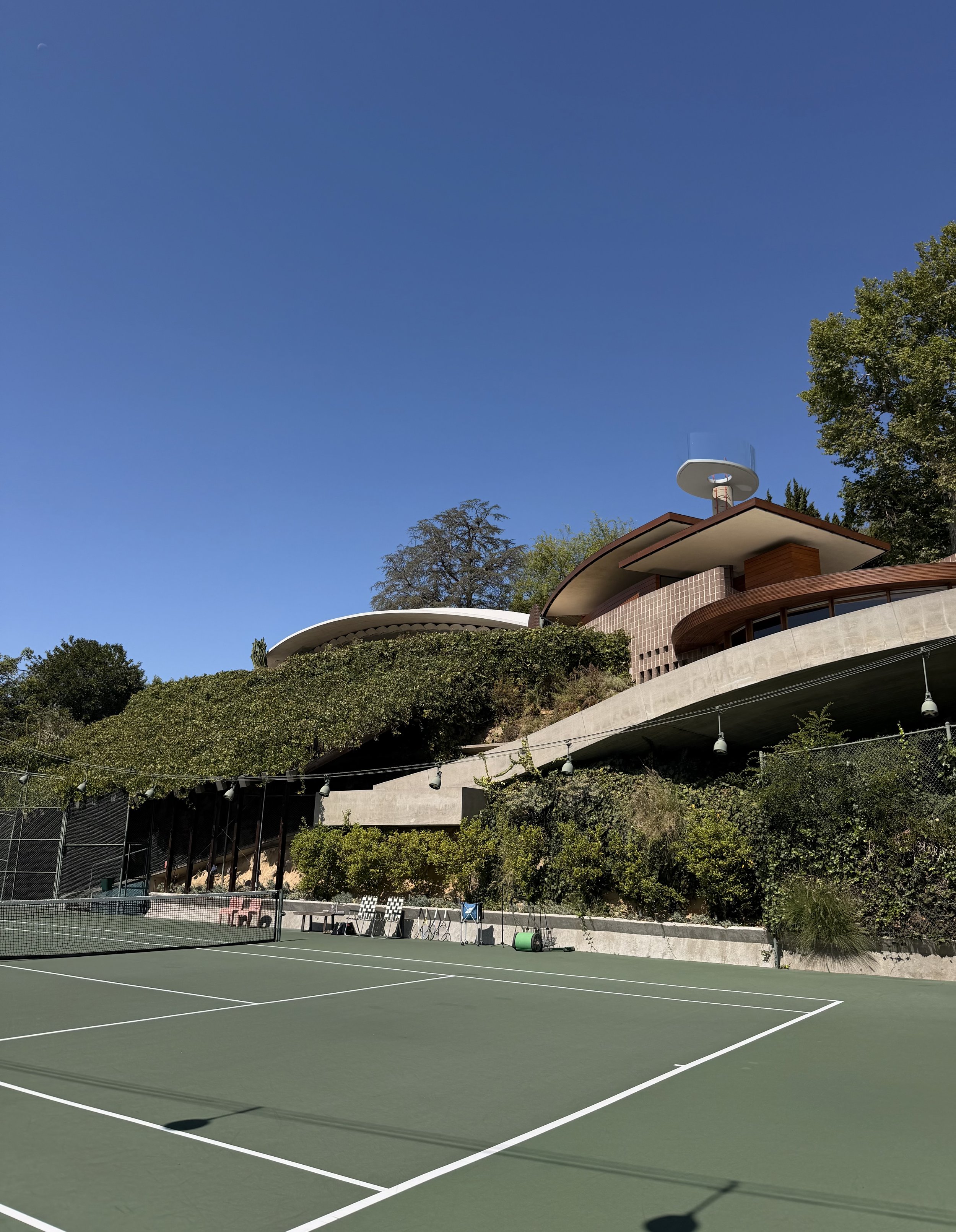 An outdoor tennis court with green surface, surrounded by trees and a modern building with a curved roof and satellite dish.