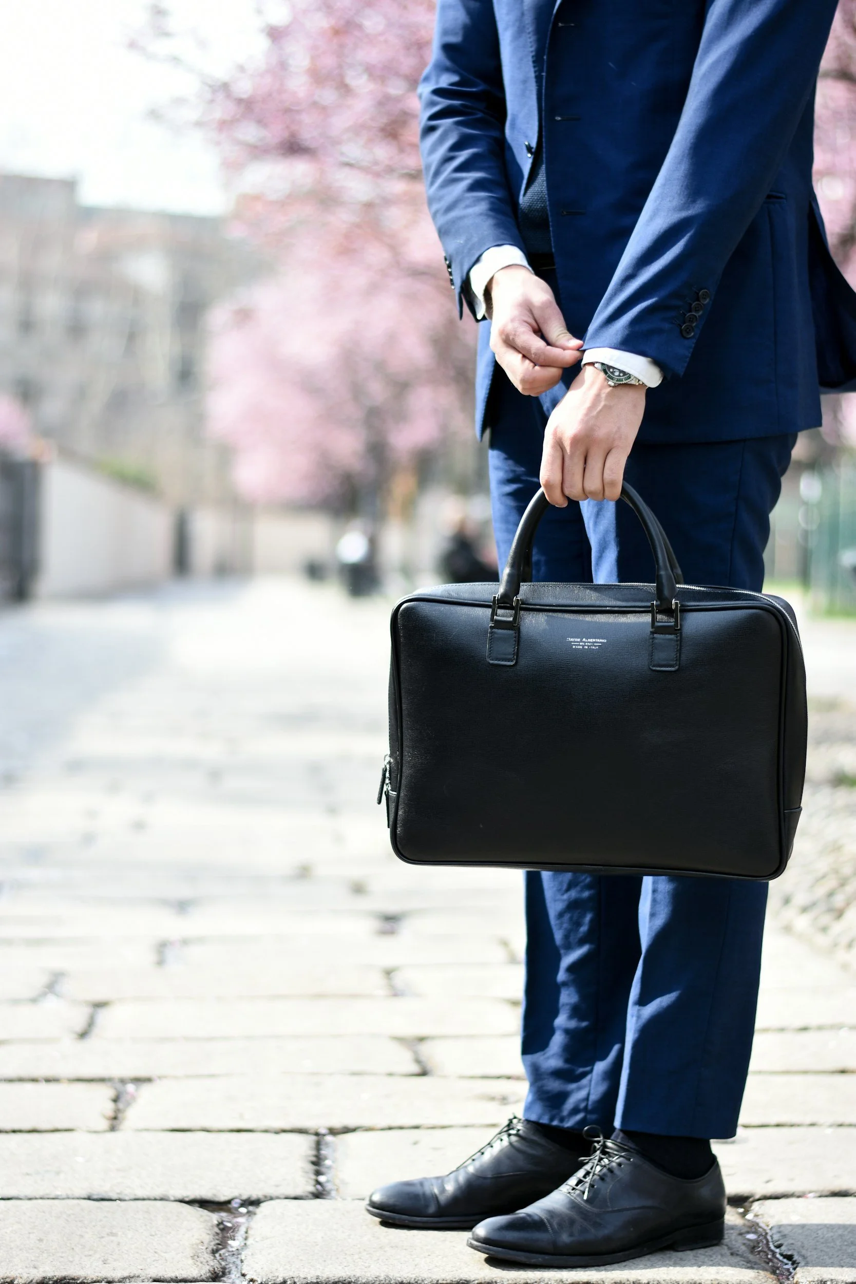 A man dressed in a navy blue suit, adjusting his shirt cuff, holding a black leather briefcase, and standing on a paved sidewalk with cherry blossom trees in the background.