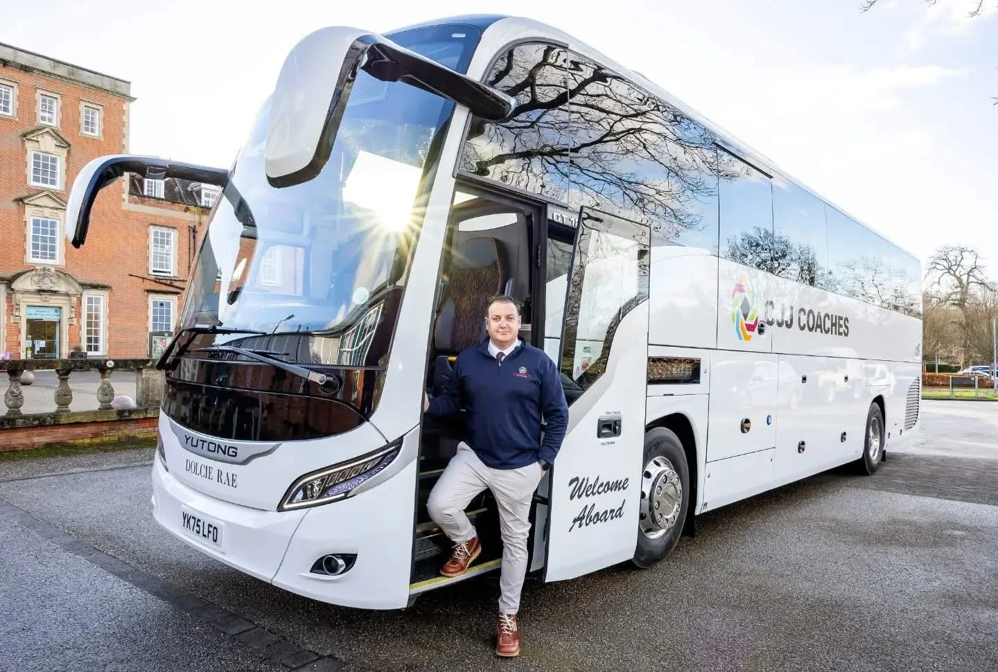 A man in a navy blue jacket and beige pants standing in the bus doorway of a white coach bus that has the words "GOJ Coaches" and "Welcome Aboard" on it, parked on a wet street with trees and brick buildings in the background.