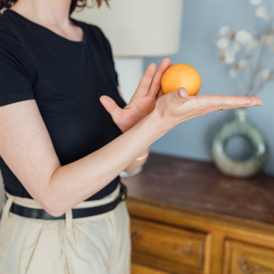 Susanna Borek hält eine Orange in ihrer Hand, im Hintergrund ein Holzkommode und eine Vase mit weißen Blumen.