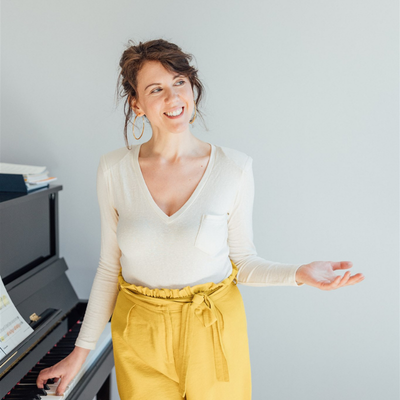 Susanna Borek mit braunen, gewellten Haaren, die in einem weißen Pullover und gelben Hosen vor einem Klavier steht, lächelnd und spricht.