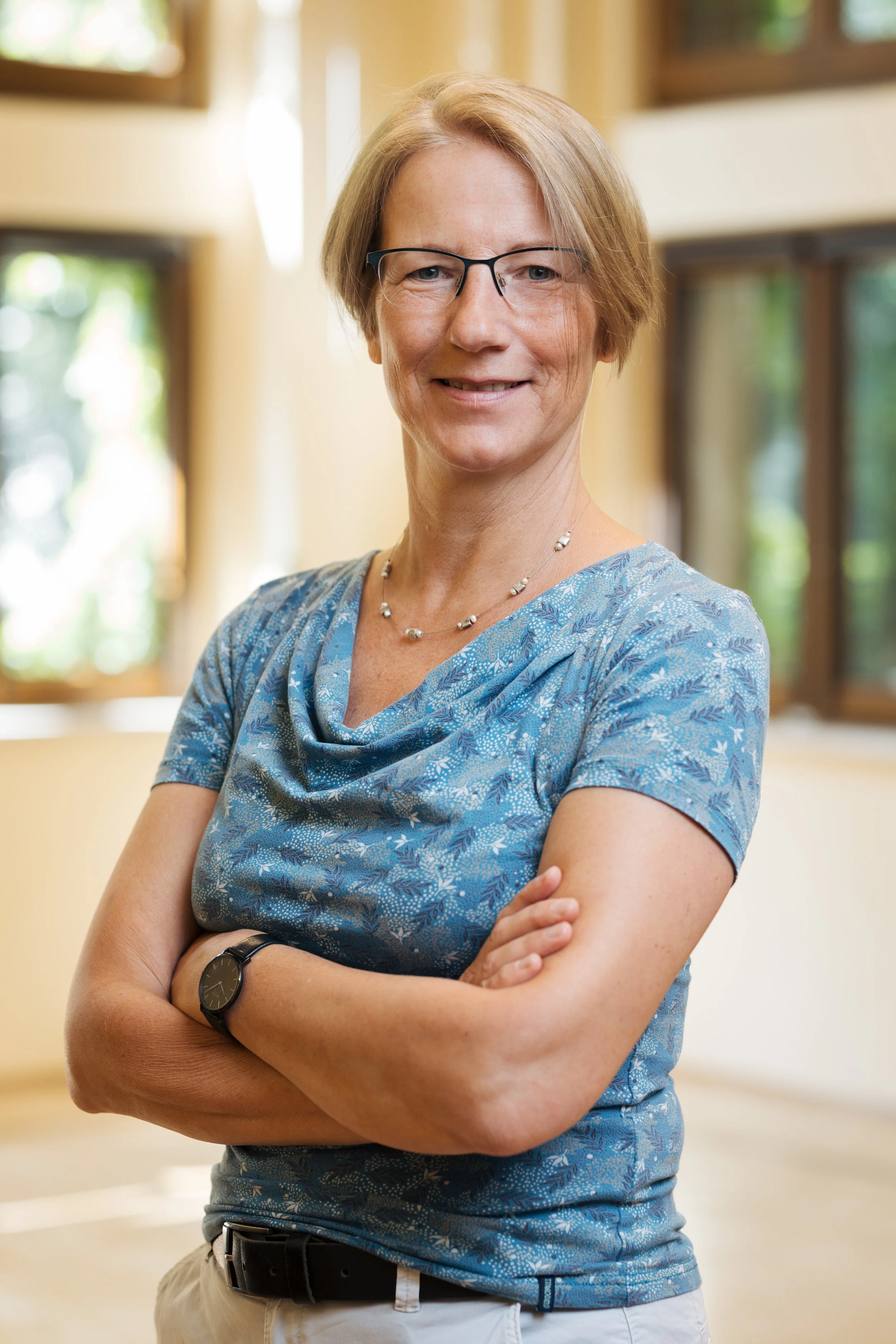 Eine Frau trägt eine blaue Bluse mit weißen Mustern, trägt eine Armbanduhr und eine Halskette, steht mit verschränkten Armen vor einem hellen Hintergrund mit Fenstern, lächelt leicht.