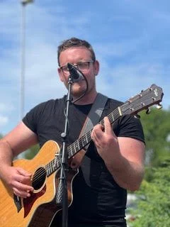 Mann spielt Gitarre und singt im Freien bei sonnigem Himmel.