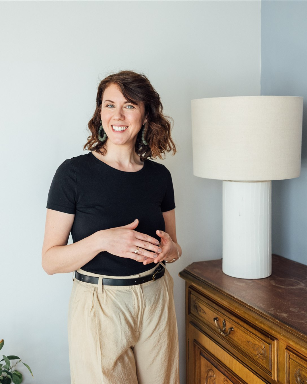 Eine lachende Frau mit braunen, lockigen Haaren trägt ein schwarzes T-Shirt und beige Hosen, steht neben einem Holztisch mit einer weißen Tischlampe.