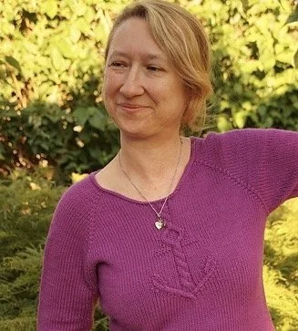 Eine lächelnde Frau mit rötlich-blondem Haar, trägt eine pinke Pullover und eine Halskette, steht vor einem grünen Busch im Freien.