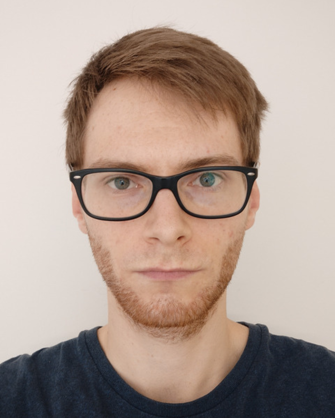 Ein junger Mann mit kurzen braunen Haaren, Brille mit schwarzen Rahmen und hellblauen Augen, trägt ein dunkelblaues T-Shirt, vor einem weißen Hintergrund.