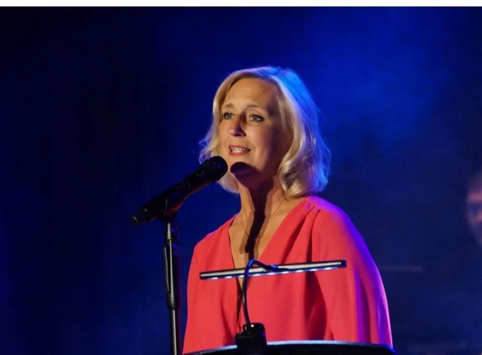 Eine blonde Frau singt bei einer Live-Performance an einem Mikrofon, im Hintergrund dunkler, blauer Lichtschein.