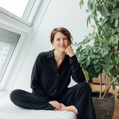 Susanna Borek sitzt auf einem Bett neben einer großen Topfpflanze vor einem Fenster.