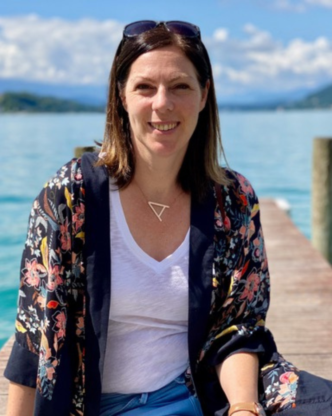 Frau sitzt auf einem Steg am Wasser, trägt eine Sonnenbrille auf dem Kopf, ein weißes T-Shirt, eine dunkle Jacke mit Blumenmuster, eine Halskette mit einem geometrischen Anhänger und lächelt. Im Hintergrund sind Wasser, Himmel mit Wolken und Berge sichtbar.