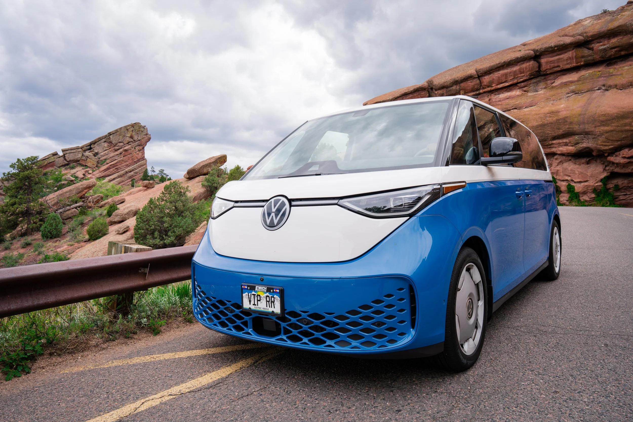Blue and white Volkswagen electric vehicle parked on a mountain road with red rock formations and cloudy sky in the background.